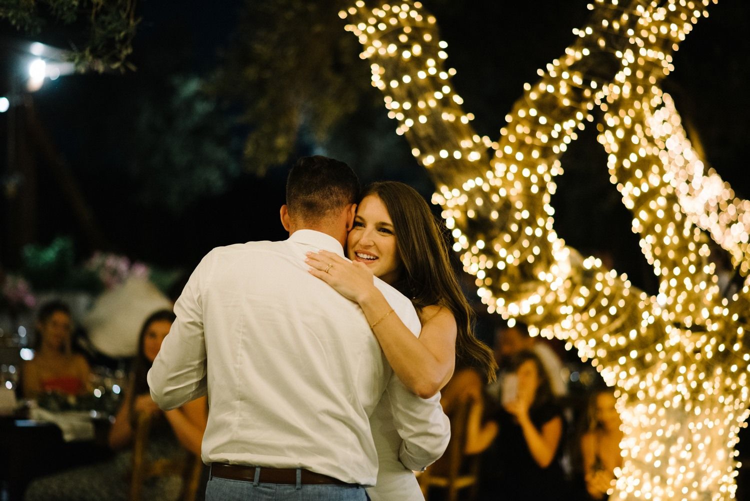 Romantic moment captured under trees wrapped in glowing fairy lights at evening wedding reception.