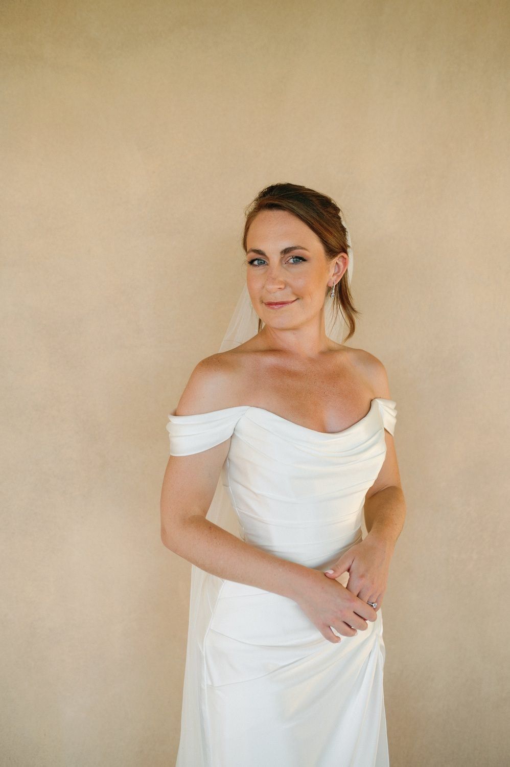 A bride poses in an off-shoulder white wedding gown against a neutral beige wall.