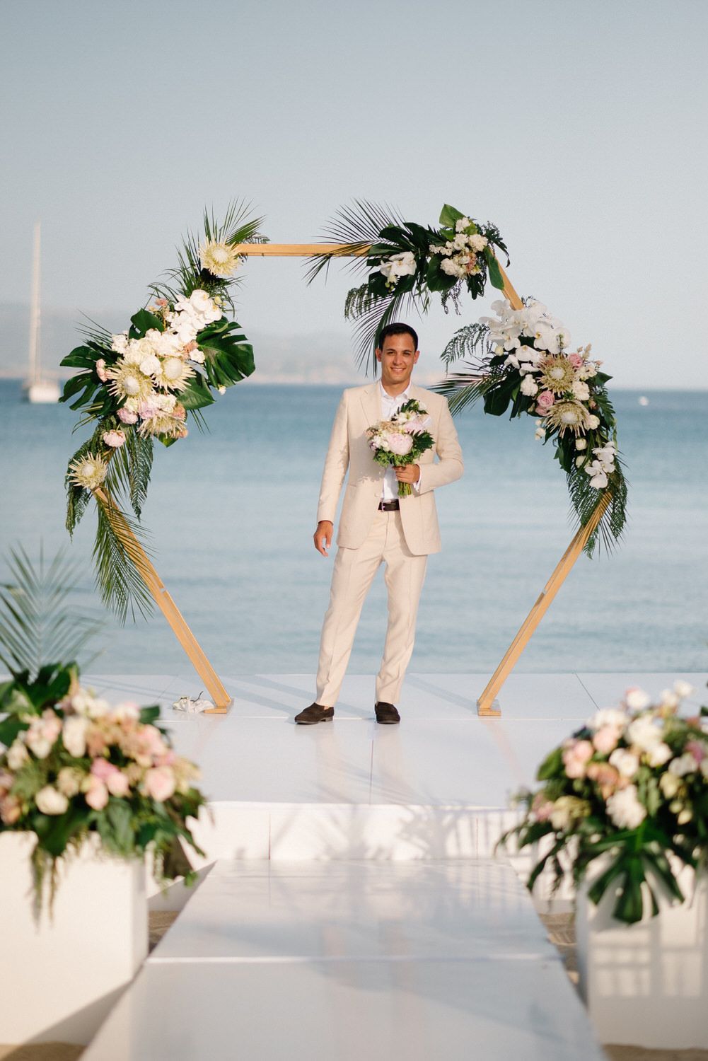 Elegant beachfront wedding ceremony setup with a hexagonal arch adorned with tropical flowers and white blooms.