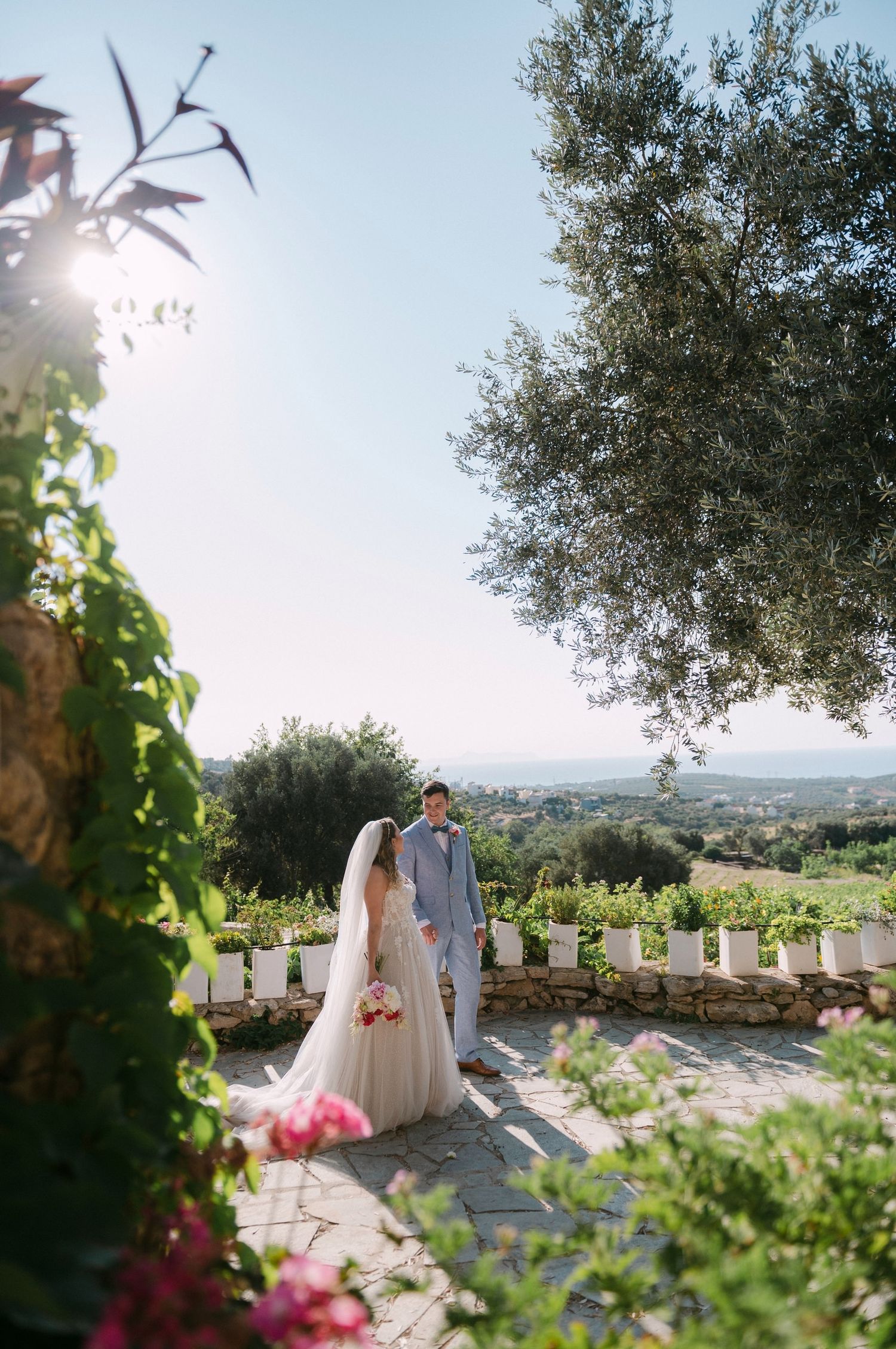 A wedding couple stands in a scenic Mediterranean garden terrace overlooking a coastal landscape on a sunny day.
