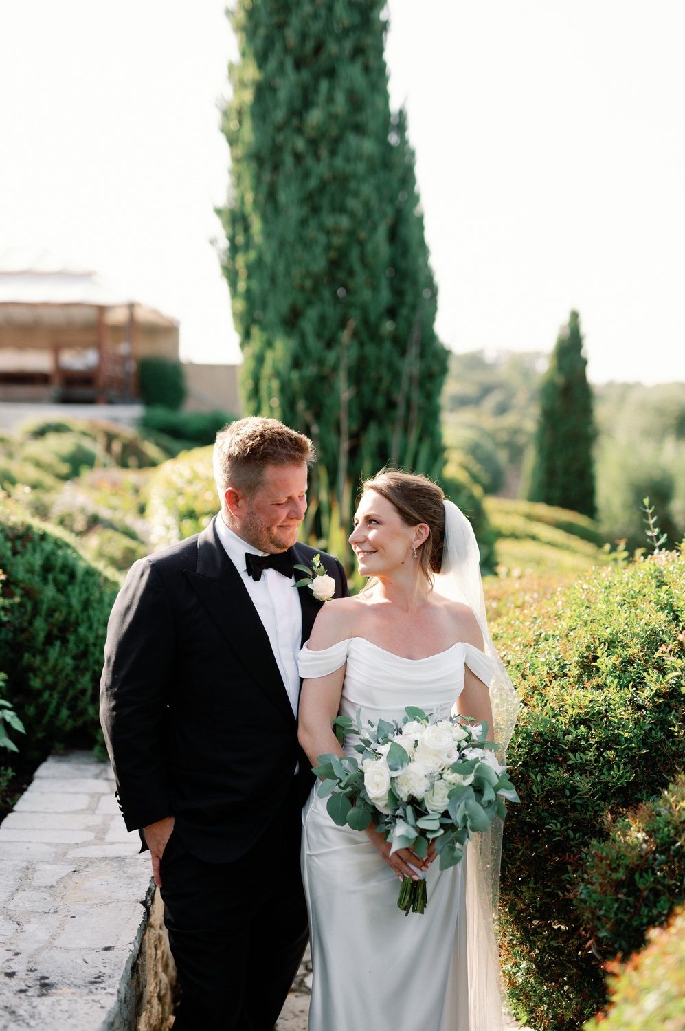 Elegant wedding couple poses in lush Mediterranean garden with cypress trees.