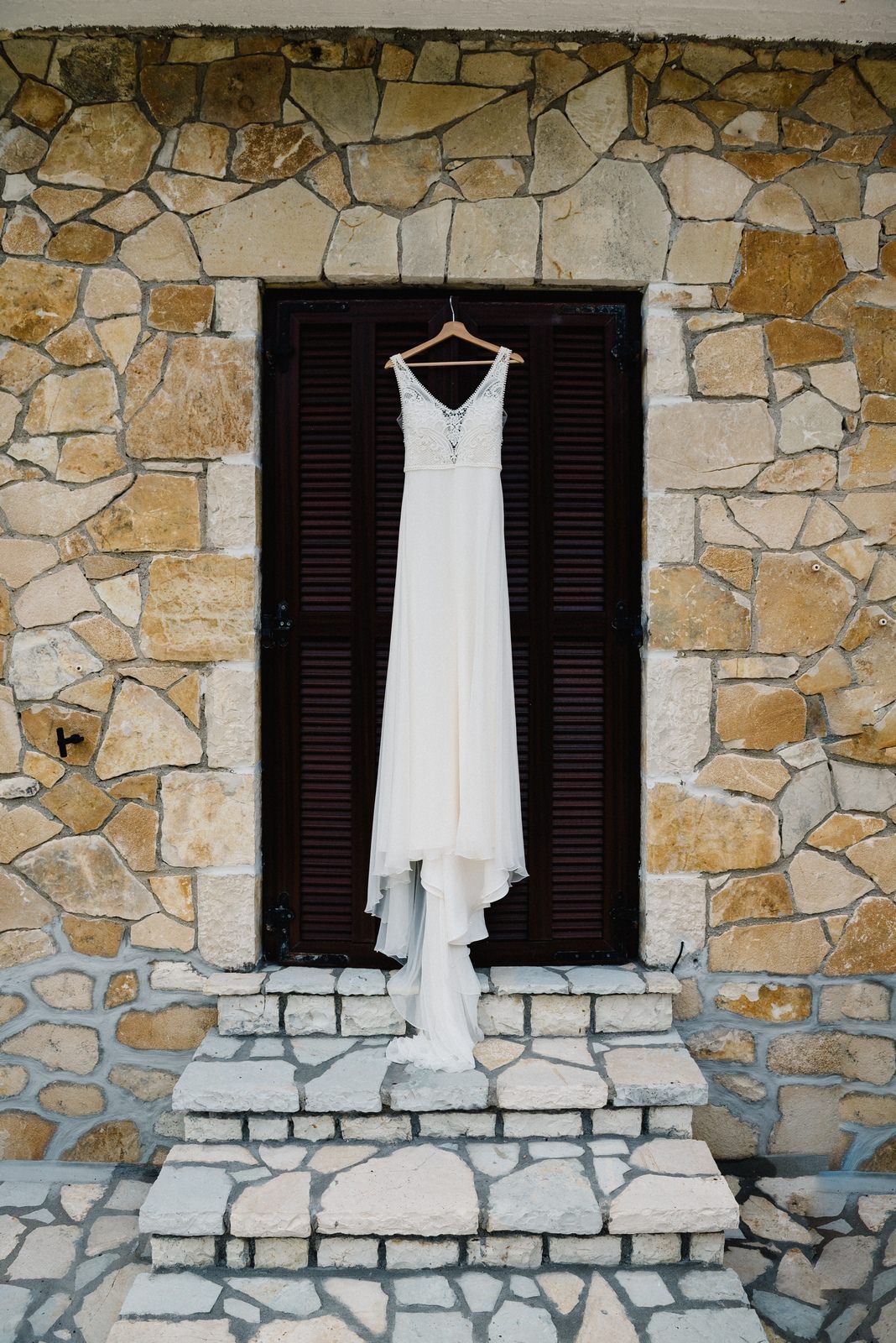 White wedding dress hanging on stone wall with dark shutters creating dramatic contrast.