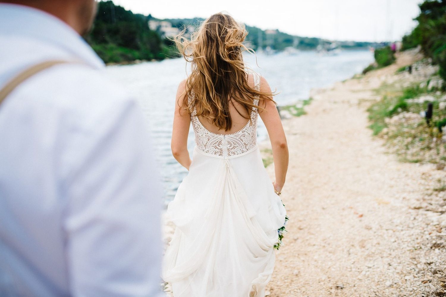 A bride in a lace dress walking along a coastal path with water views.