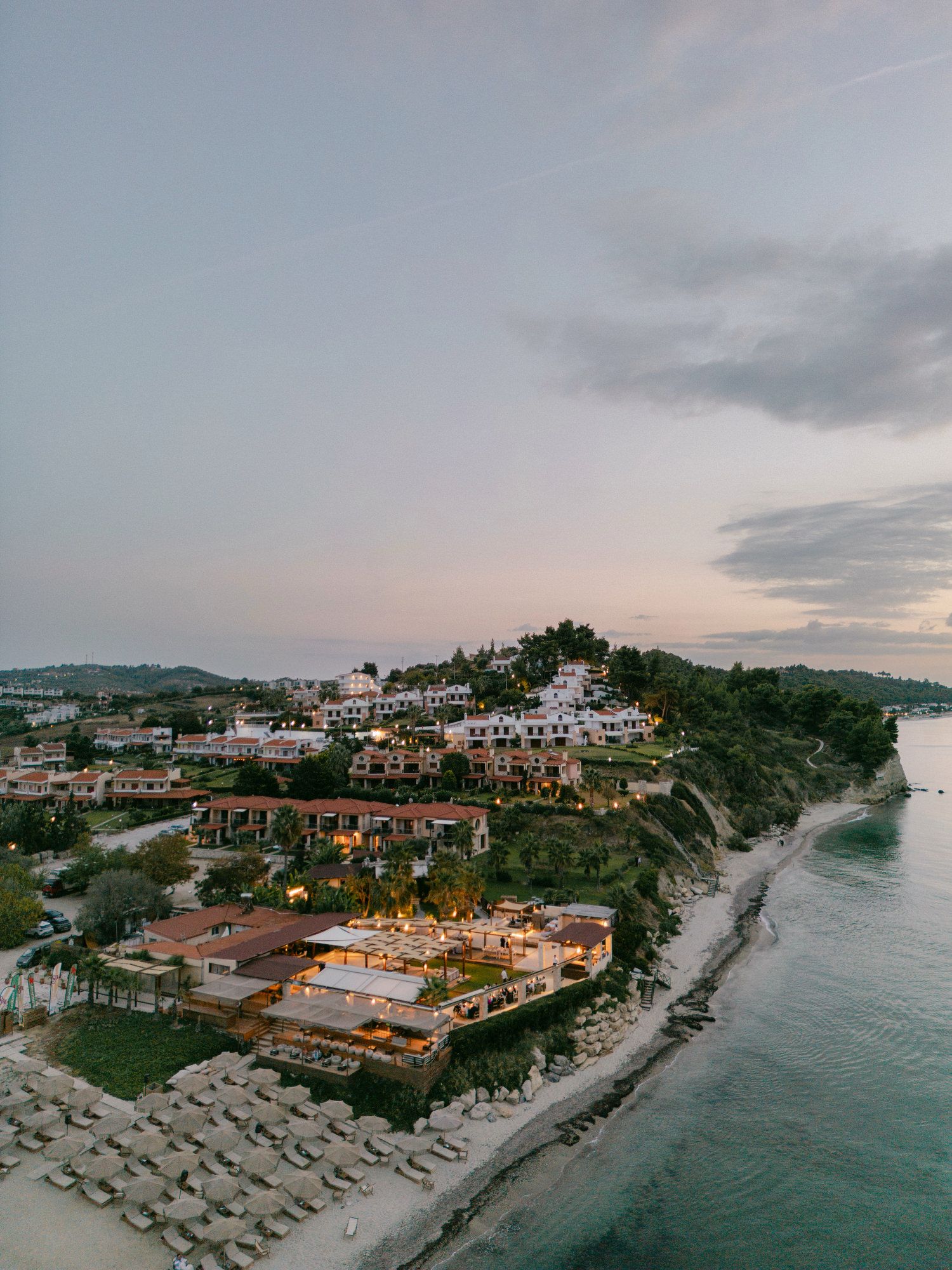 Aerial panoramic view of a coastal resort with beach umbrellas and Mediterranean-style buildings at sunset.