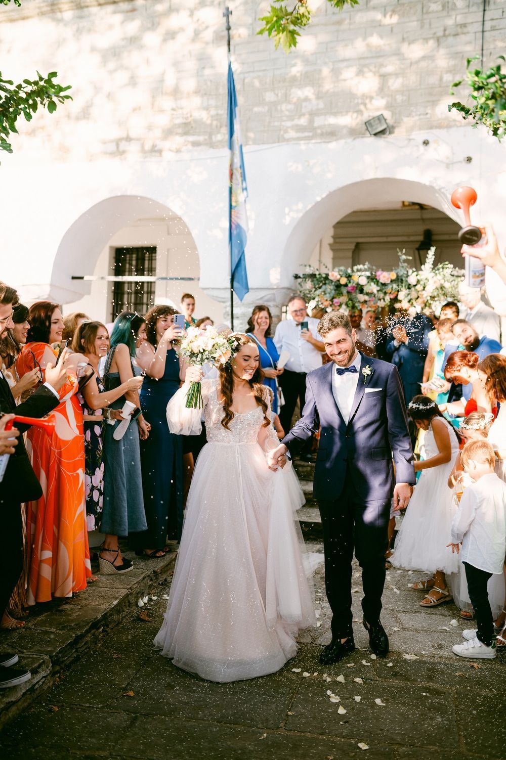 A wedding celebration sequence showing guests gathered outside a white stone church in Pelion, Greece
