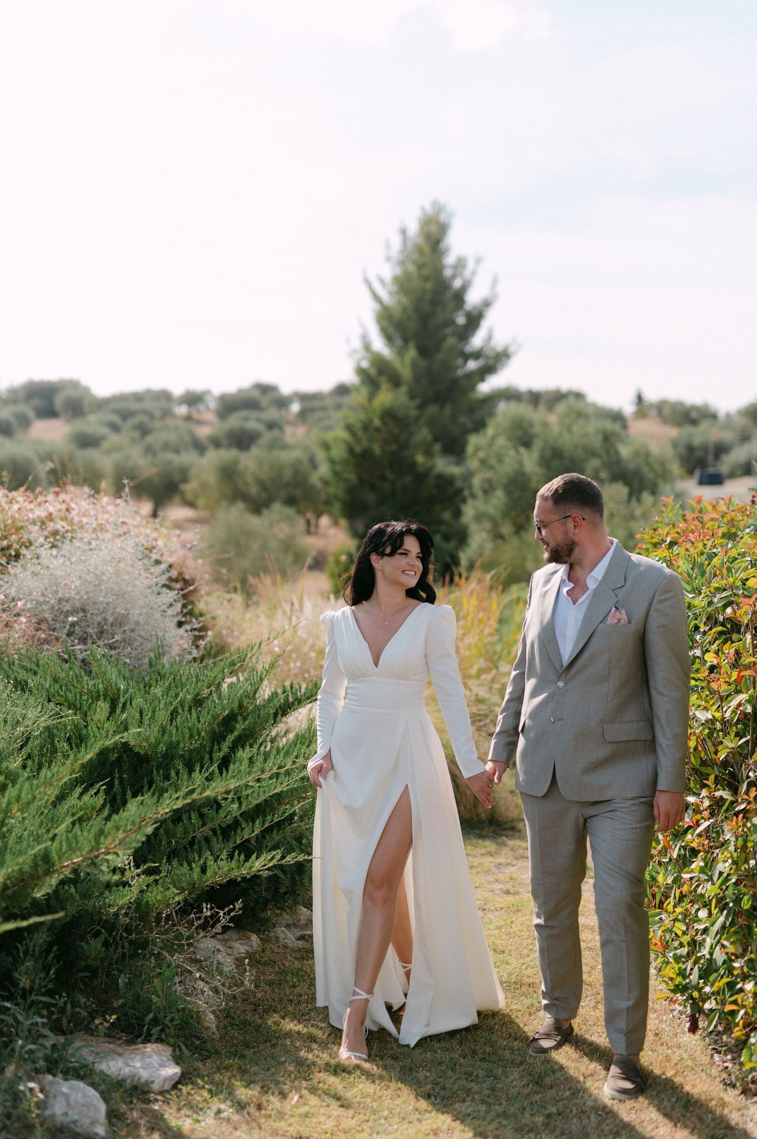 A couple walking hand in hand through a garden path surrounded by greenery and wildflowers on their wedding day.