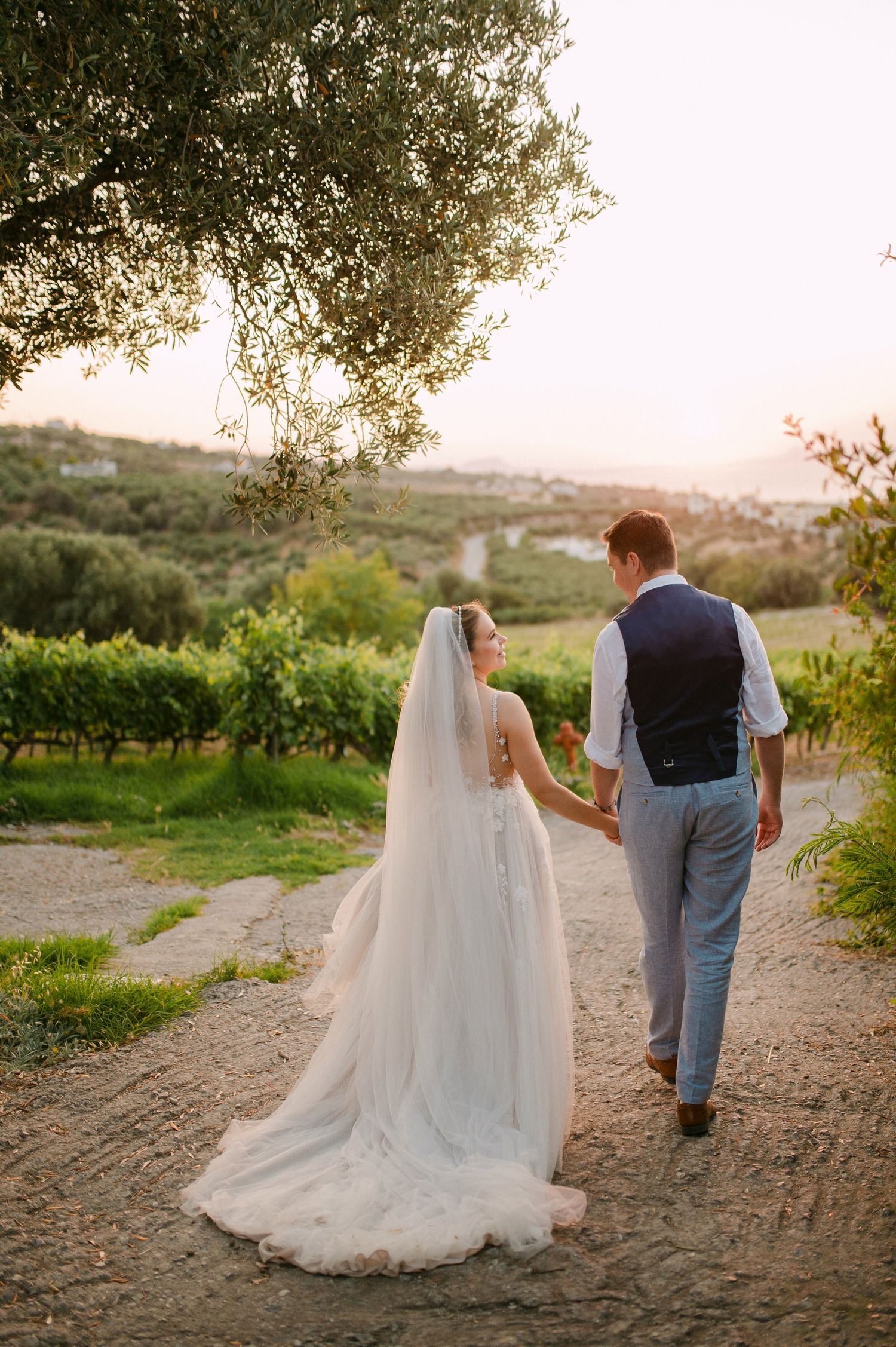 A wedding couple walks hand in hand down a dirt path at sunset through a scenic vineyard landscape.