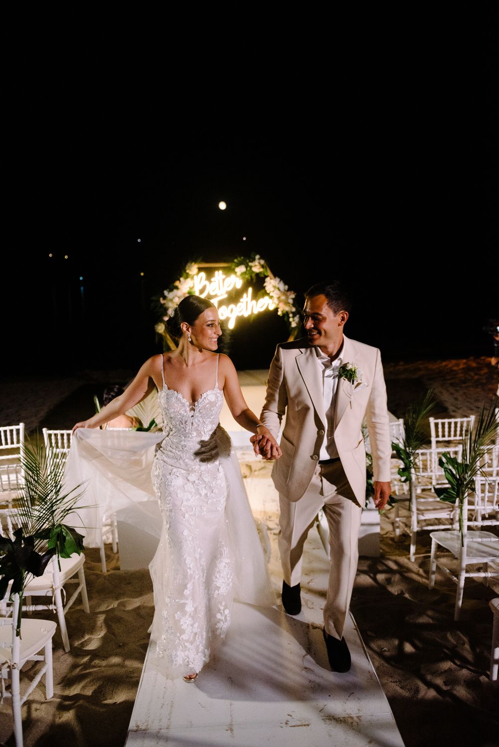 A couple walks hand in hand at a nighttime beach wedding ceremony with illuminated decor and white chairs.