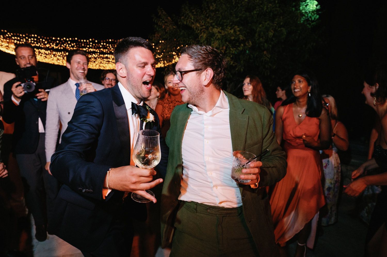 Two party guests sharing a lively moment while holding drinks at an outdoor evening celebration.