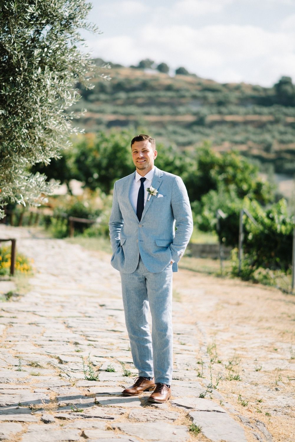 Groom in light blue suit standing on stone path with rolling hills in background.