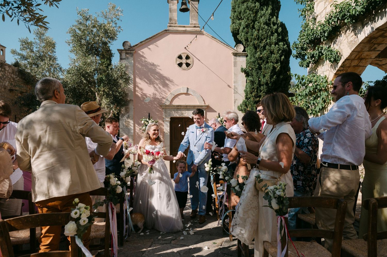 Religious ceremony taking place outside a stone church with cypress trees and pink walls in the background.