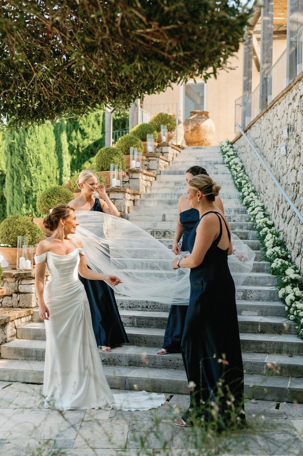 Wedding party members in navy dresses help adjust a bride's gown on stone steps lined with flowers.