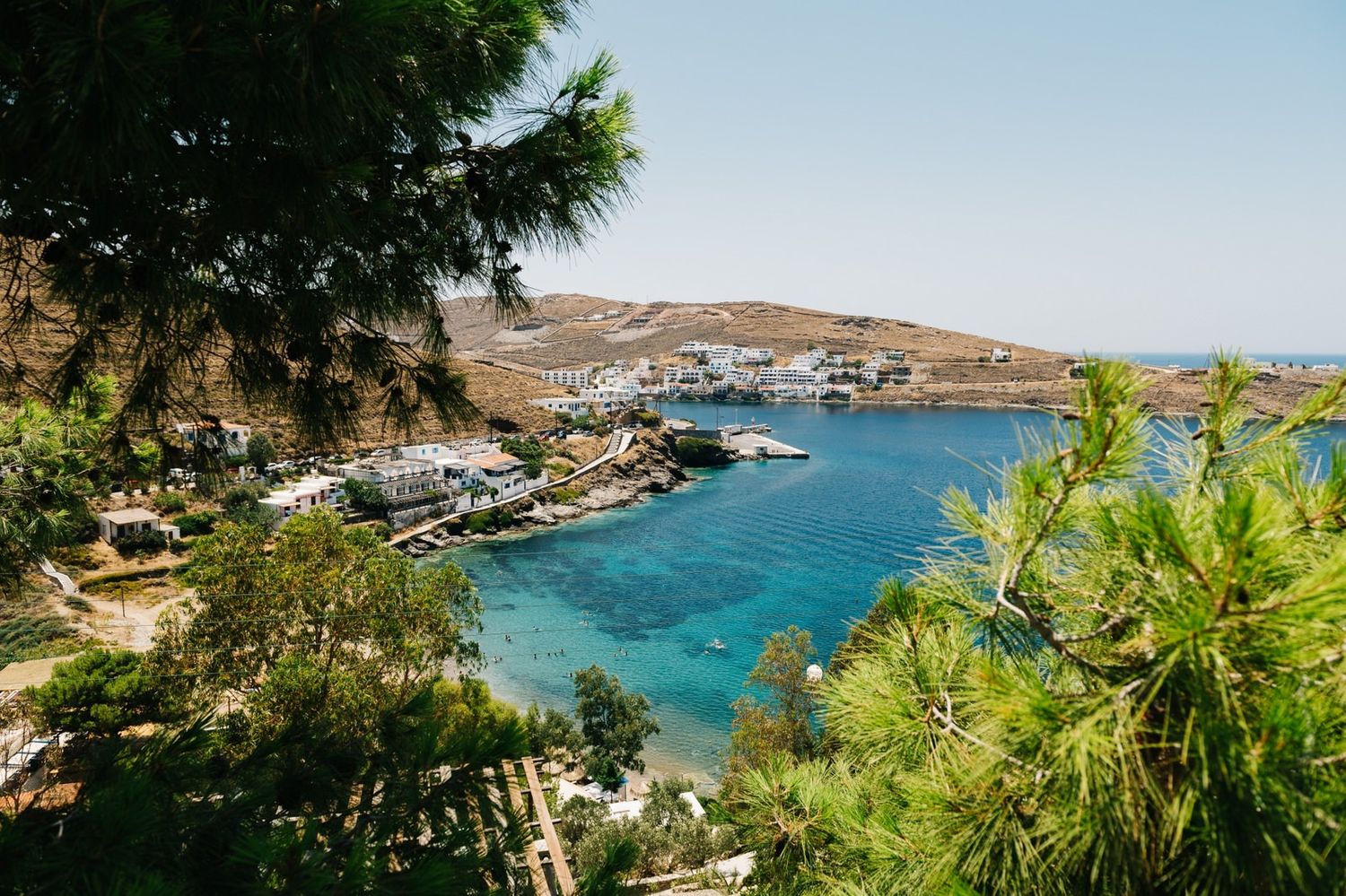 Picturesque harbor view with boats and white buildings nestled in a Mediterranean cove framed by pine trees.