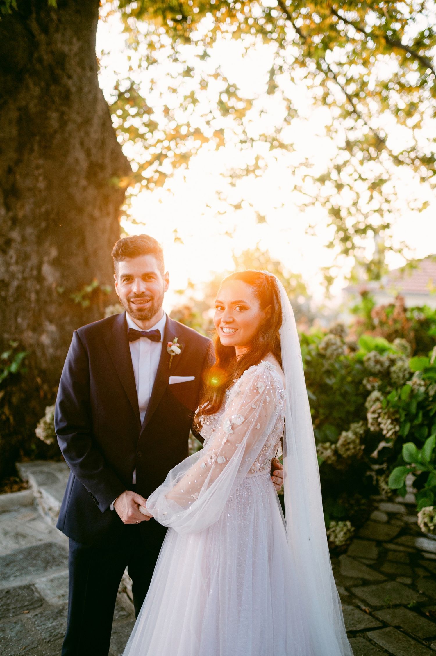 A couple in wedding attire poses together outdoors during golden hour with sunlight filtering through trees in background.