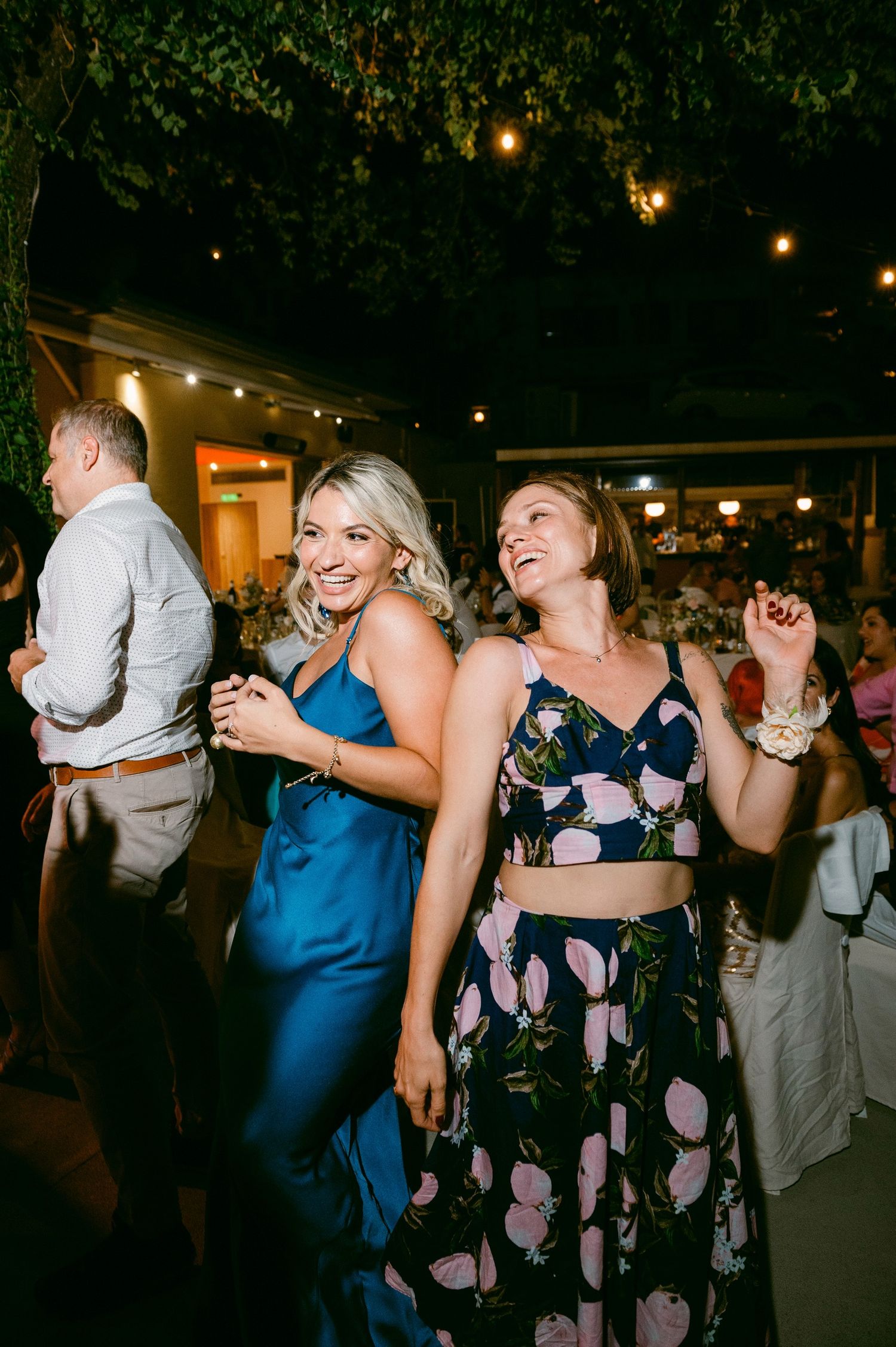 Party guests dancing at an evening celebration with string lights overhead and greenery decorations.