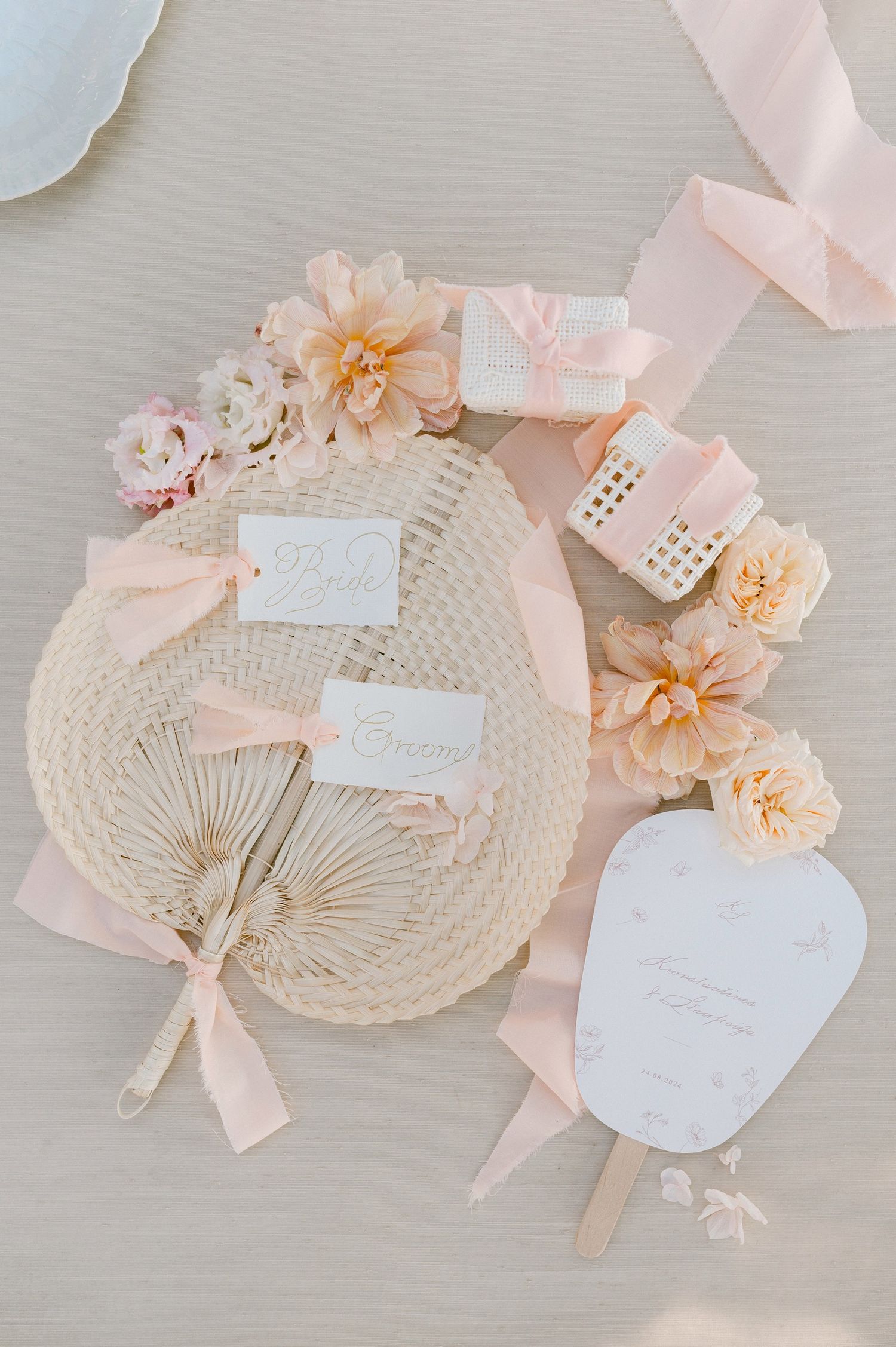Elegant wedding accessories with peach flowers, ribbons, and a woven fan arranged on a light neutral surface.