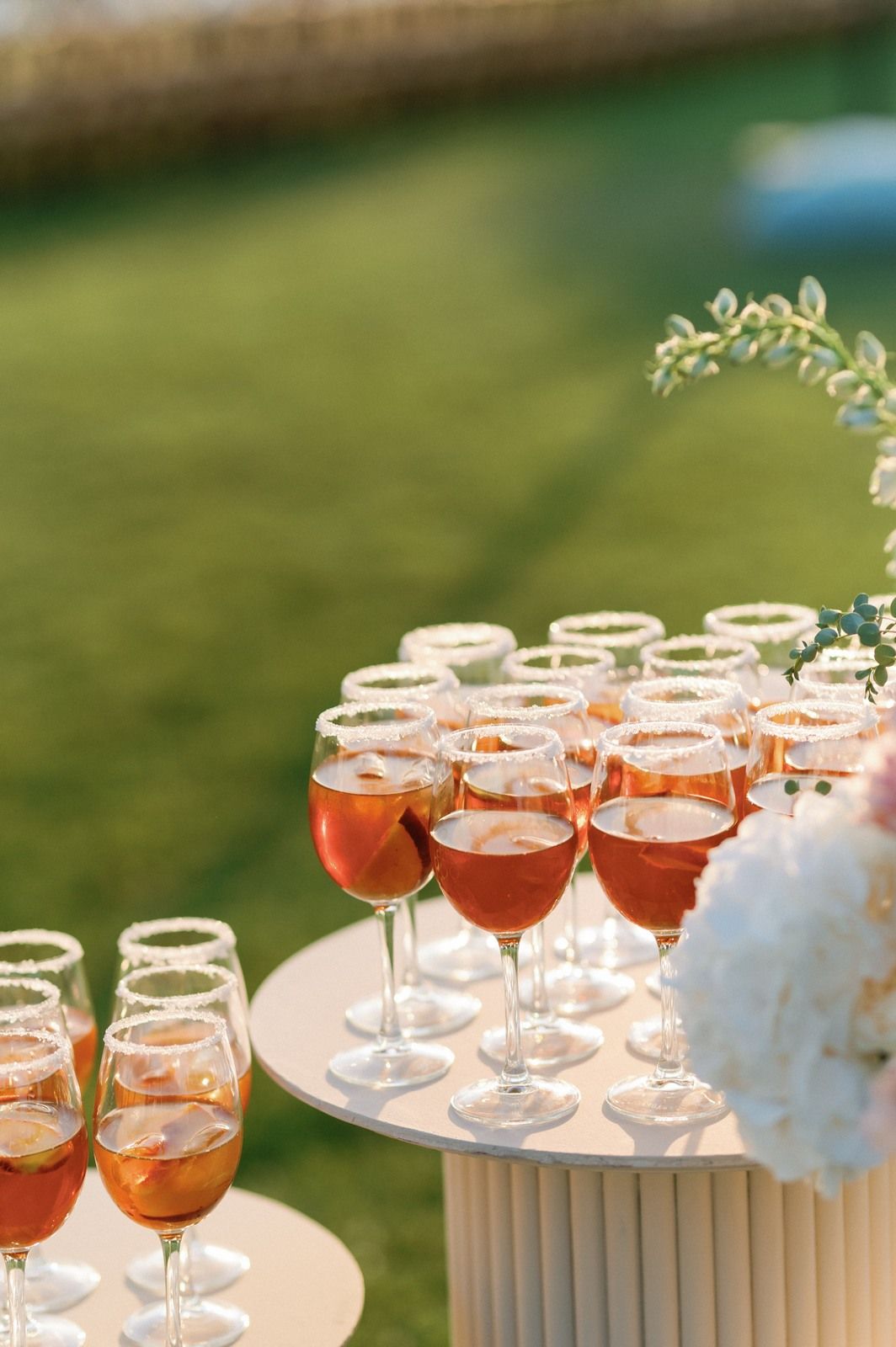 Elegant champagne glasses filled with pink cocktails arranged in rows on white tables at an outdoor garden party.