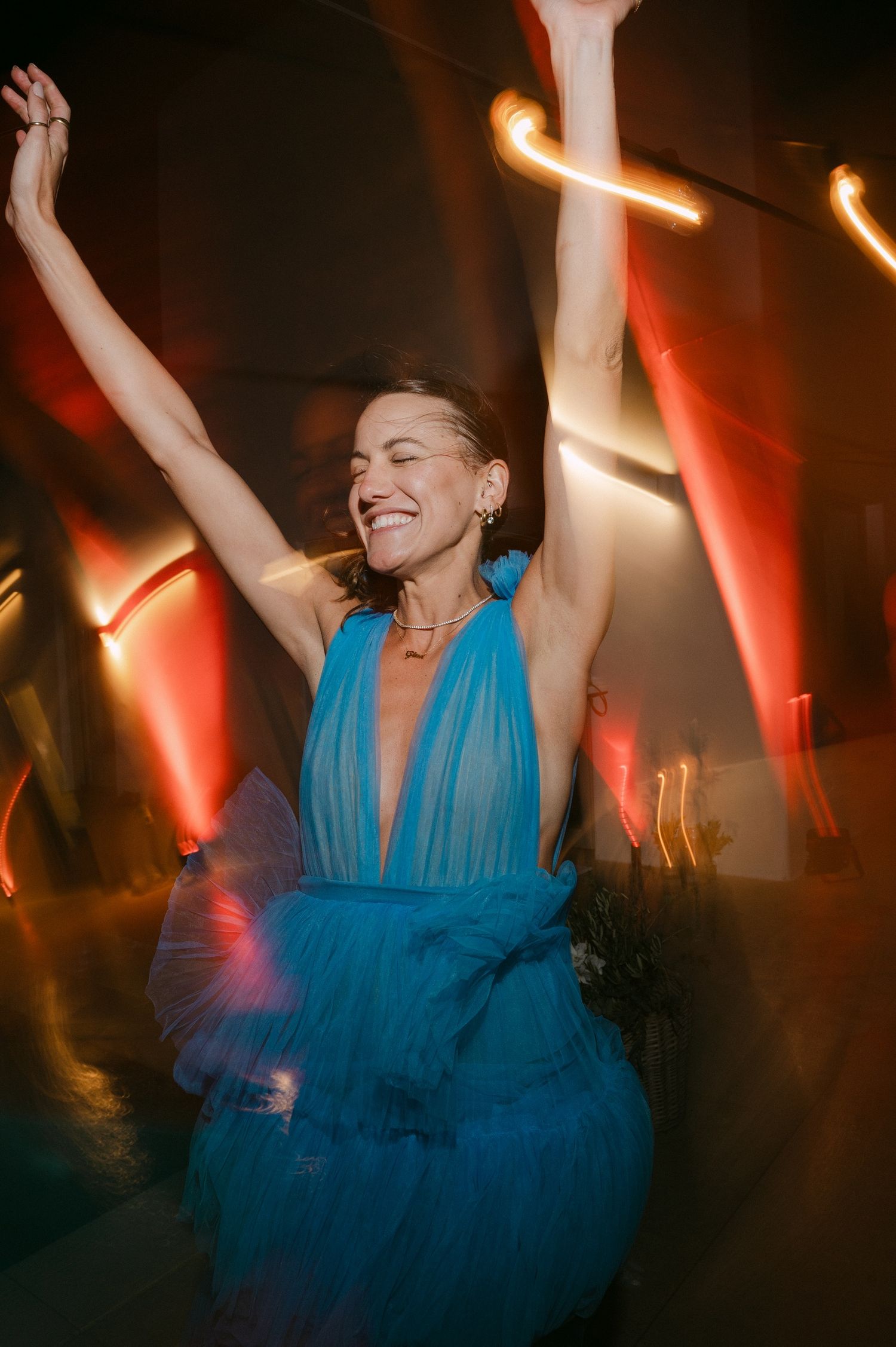 A person in a flowing turquoise dress dances joyfully with raised arms under dramatic stage lighting at a party.