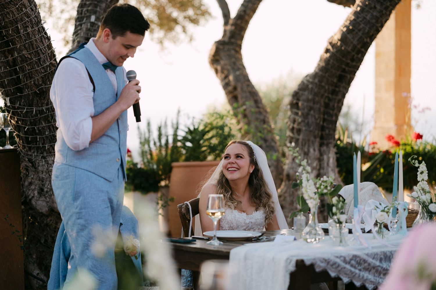 A joyful moment at an outdoor wedding reception with candlelit table settings under trees at dusk.