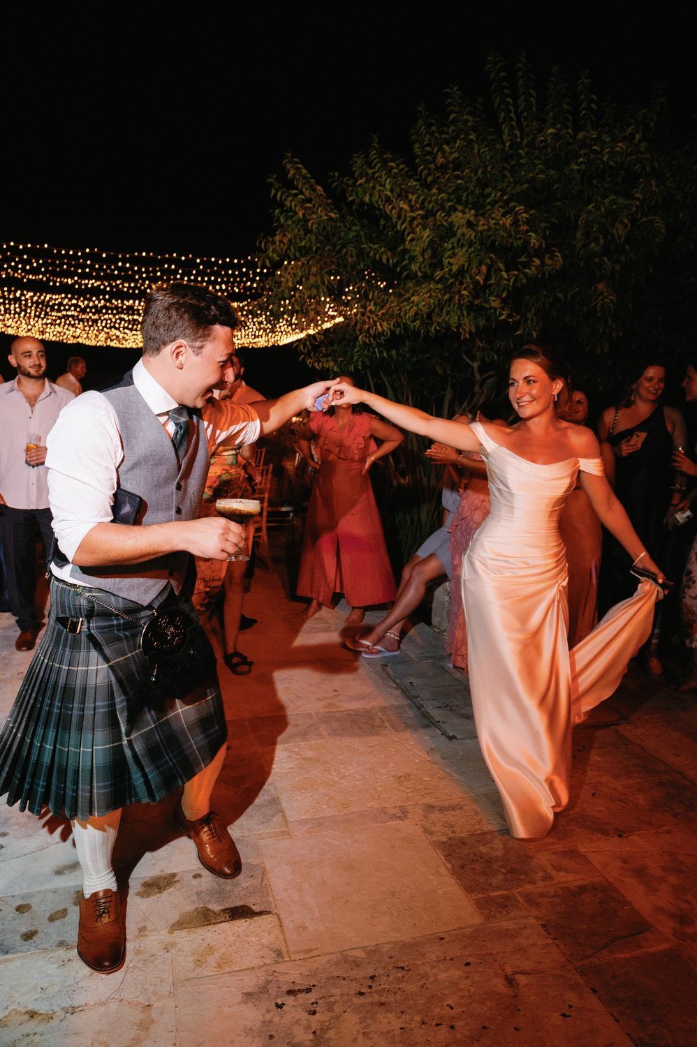 Wedding couple dancing with guests, groom wearing traditional Scottish kilt at outdoor reception.
