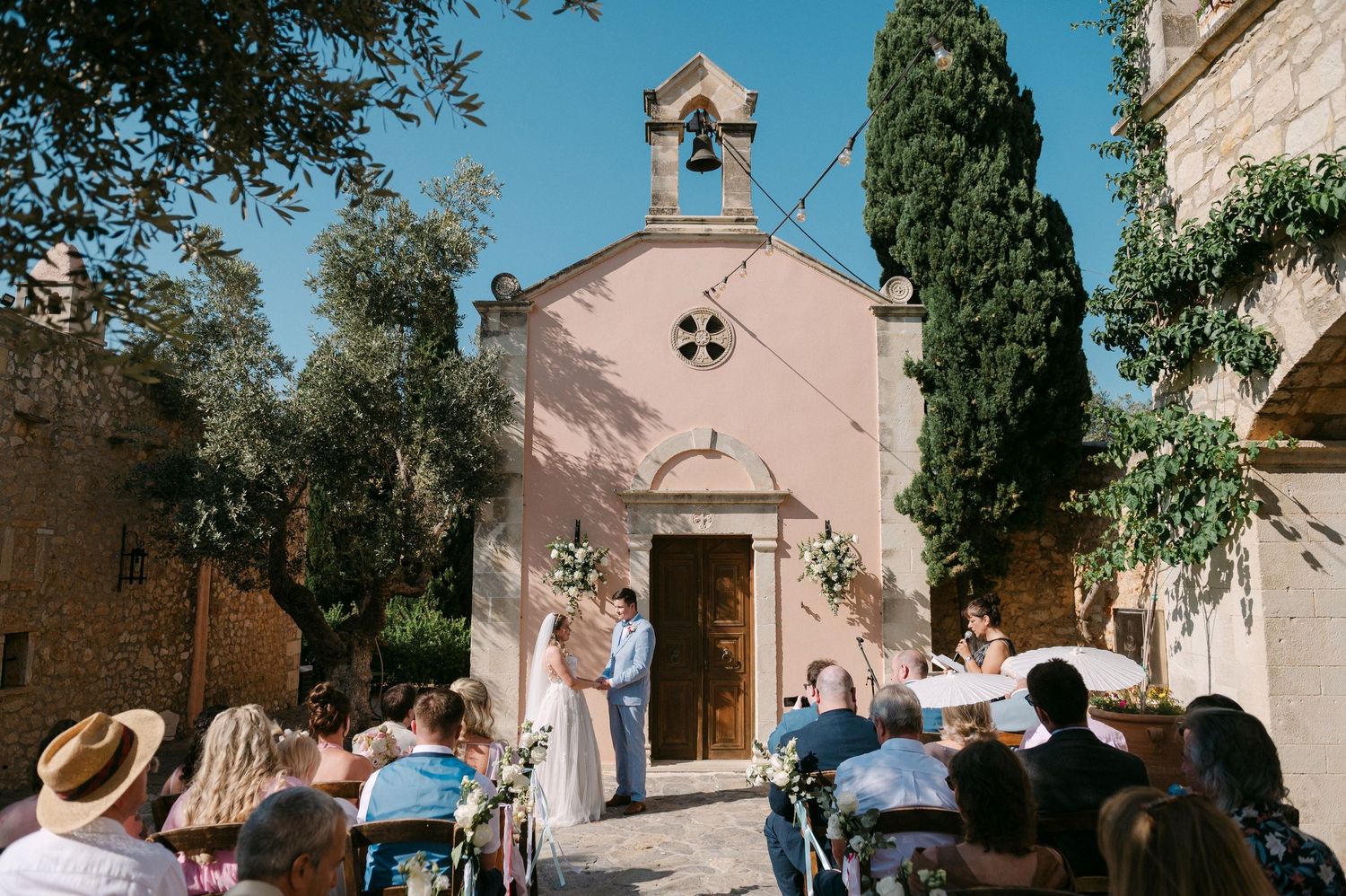 Small Mediterranean chapel with pink walls and cypress trees hosts an intimate outdoor wedding ceremony in warm sunlight.
