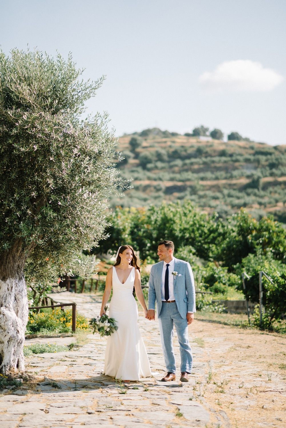 A couple walking together on a rustic stone path surrounded by olive trees and Mediterranean landscape on a sunny day.