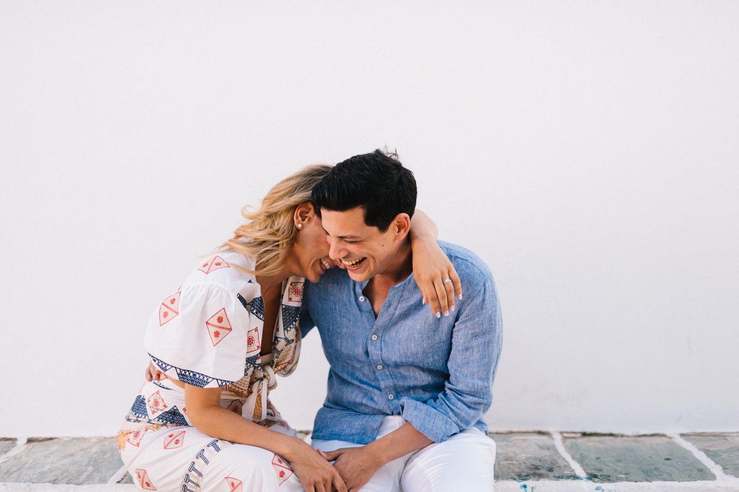 A romantic couple shares an intimate moment while sitting together against a white wall.