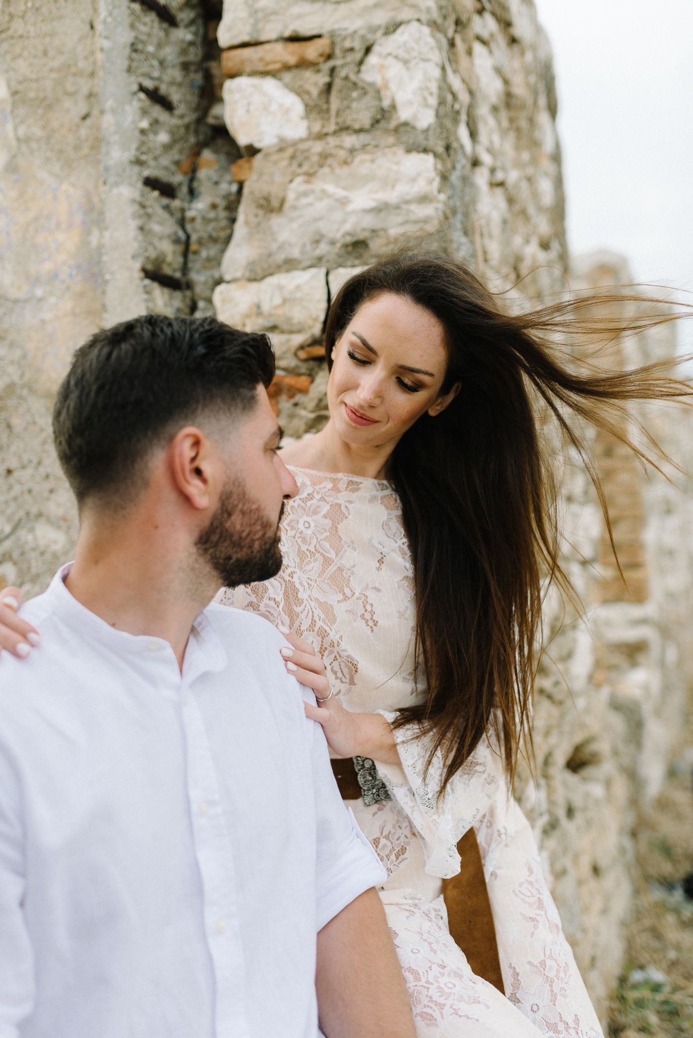 Candid moment captured with wind blowing hair against stone wall backdrop.