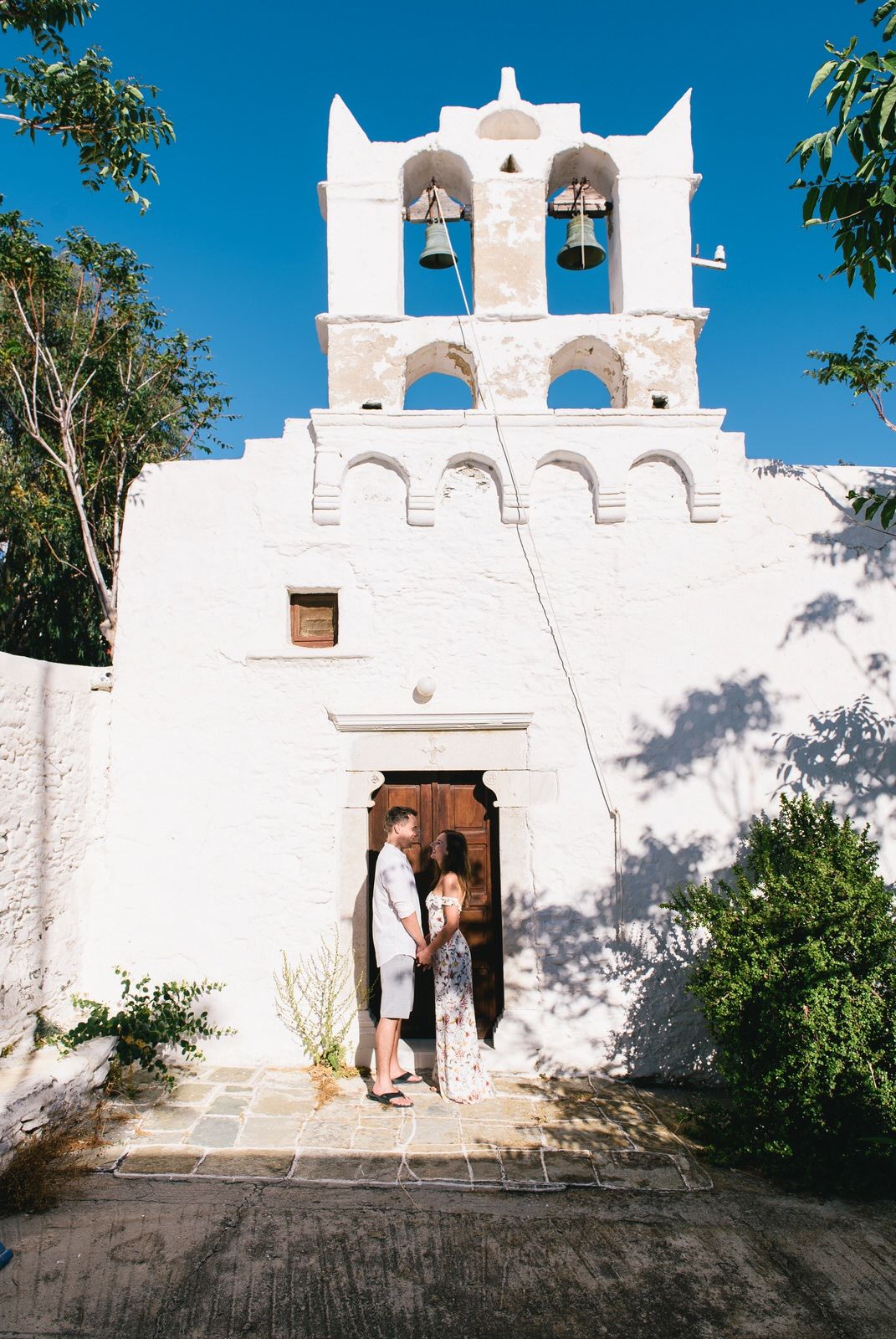 Couple standing in doorway of traditional white Greek Orthodox church with bell tower.