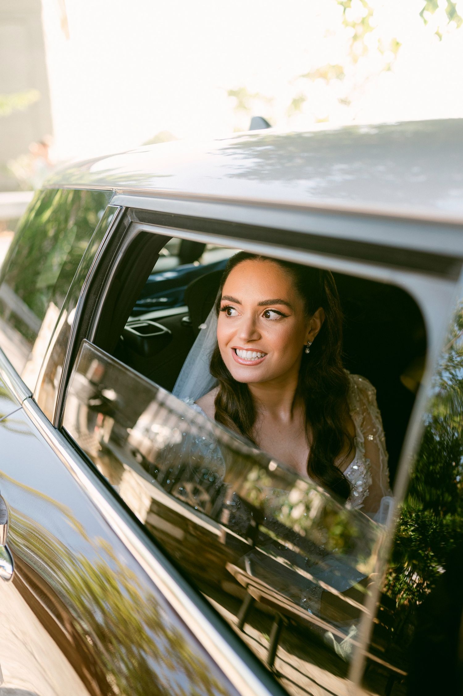 Smiling passenger looks through car window on a sunny day with sunlight creating warm reflective highlights.