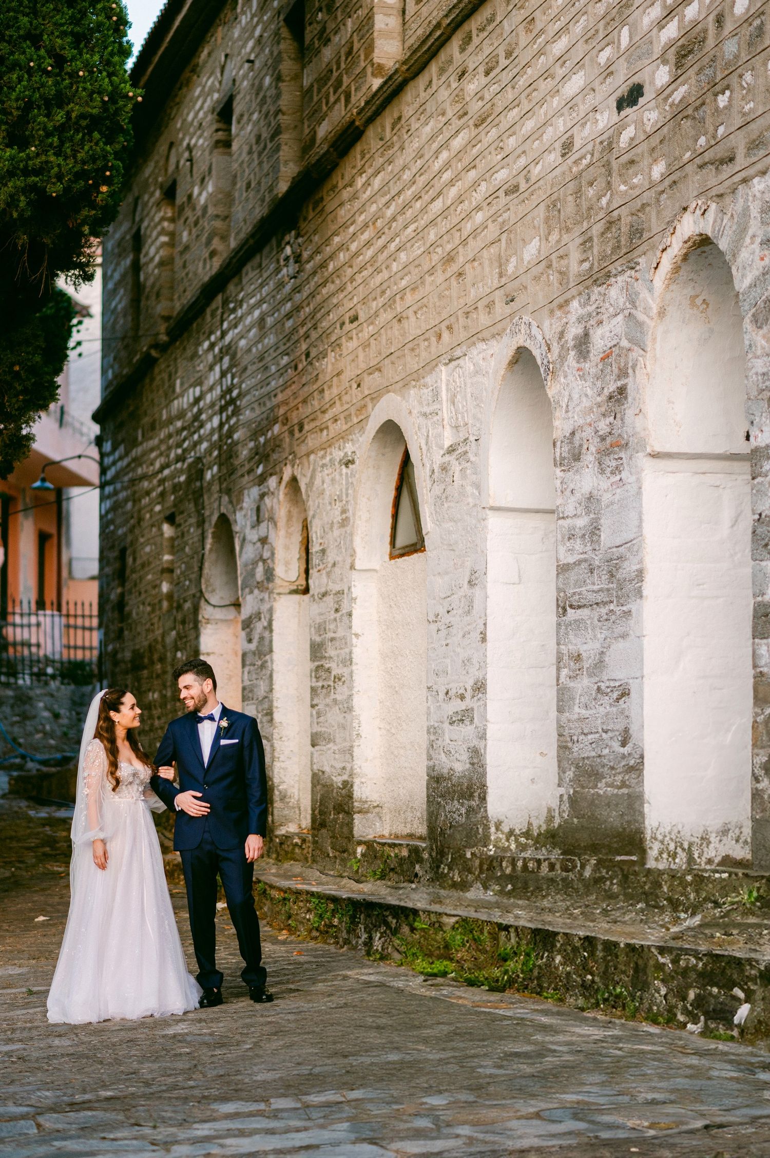 A couple poses against a historic stone wall with white archways and cobblestone ground during a wedding photo shoot.
