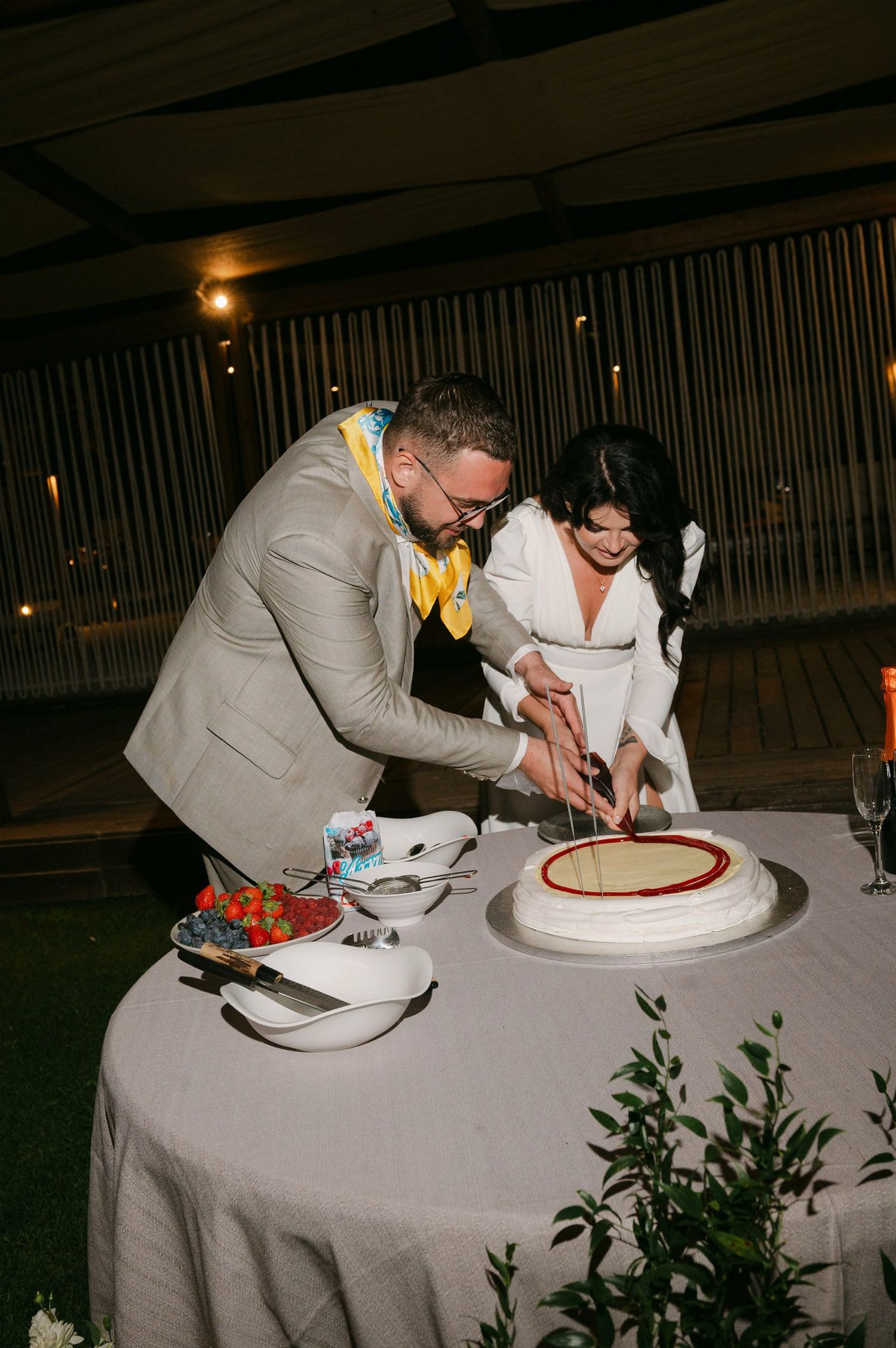 A couple cuts their wedding cake together at a reception table decorated with greenery and white linens.