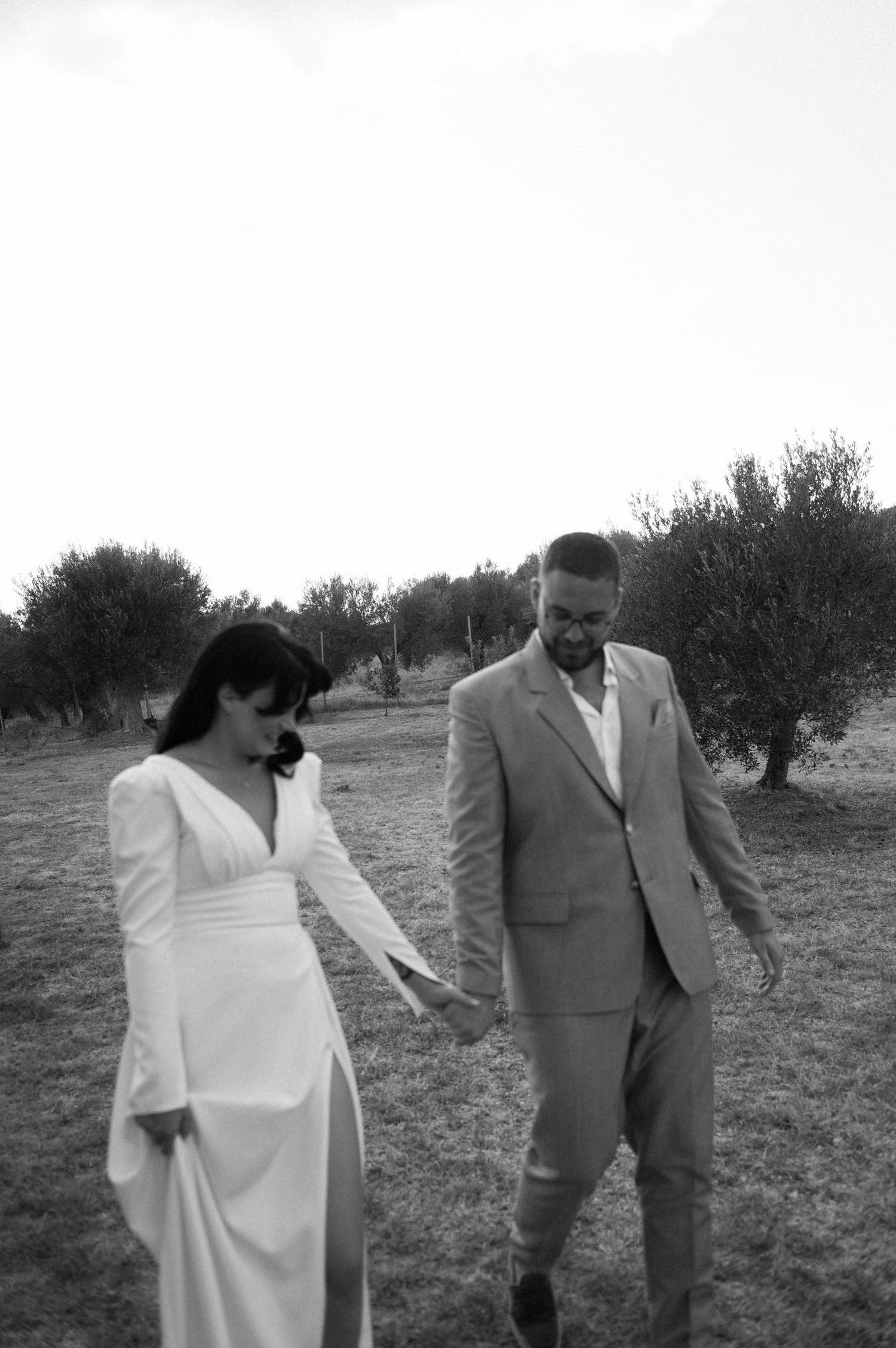 A couple in wedding attire poses together in a rustic outdoor setting with olive trees in the background.