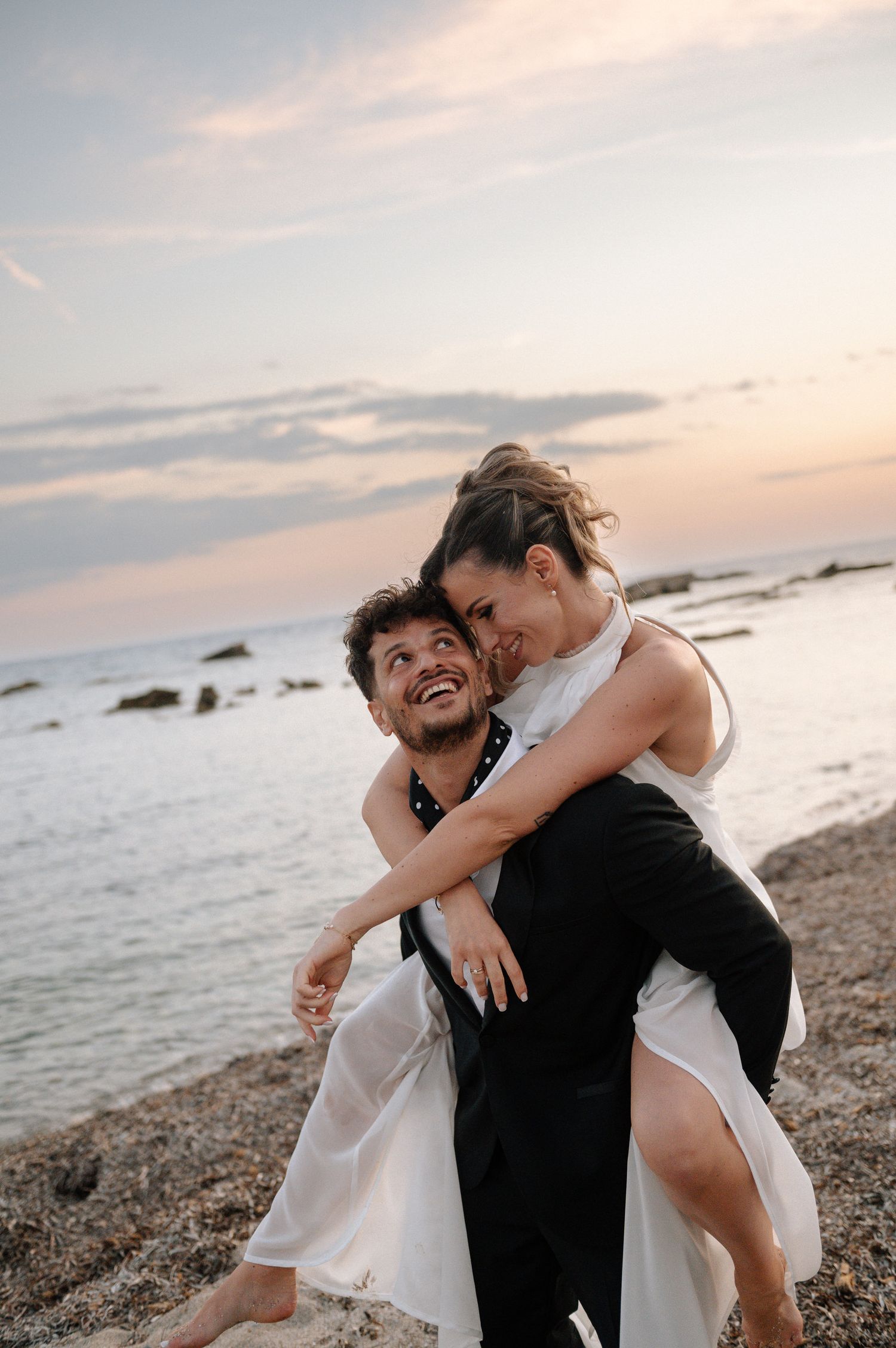 A joyful couple in wedding attire sharing a piggyback ride on a beach at sunset.