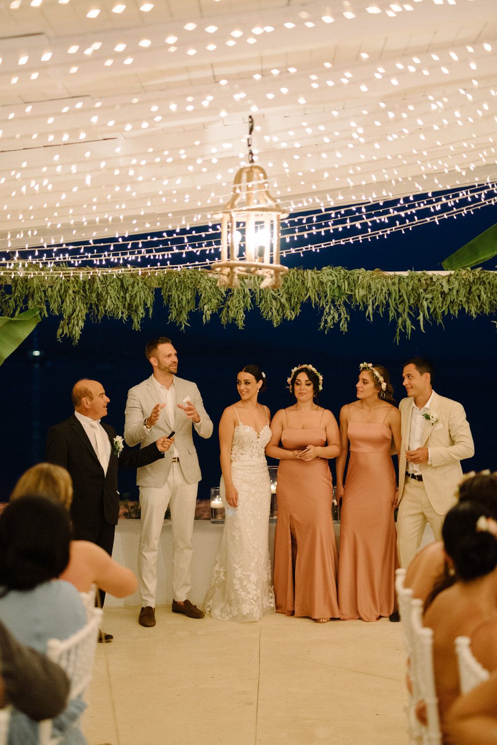 Wedding party members gather for speeches under a beautifully lit outdoor reception canopy.