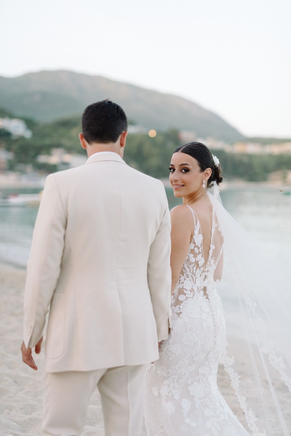 A wedding couple stands on the beach with the bride in a lace gown looking over her shoulder.