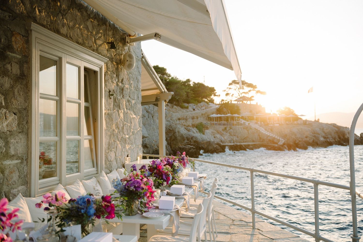Seaside restaurant terrace with elegant table settings and floral decorations at golden hour.