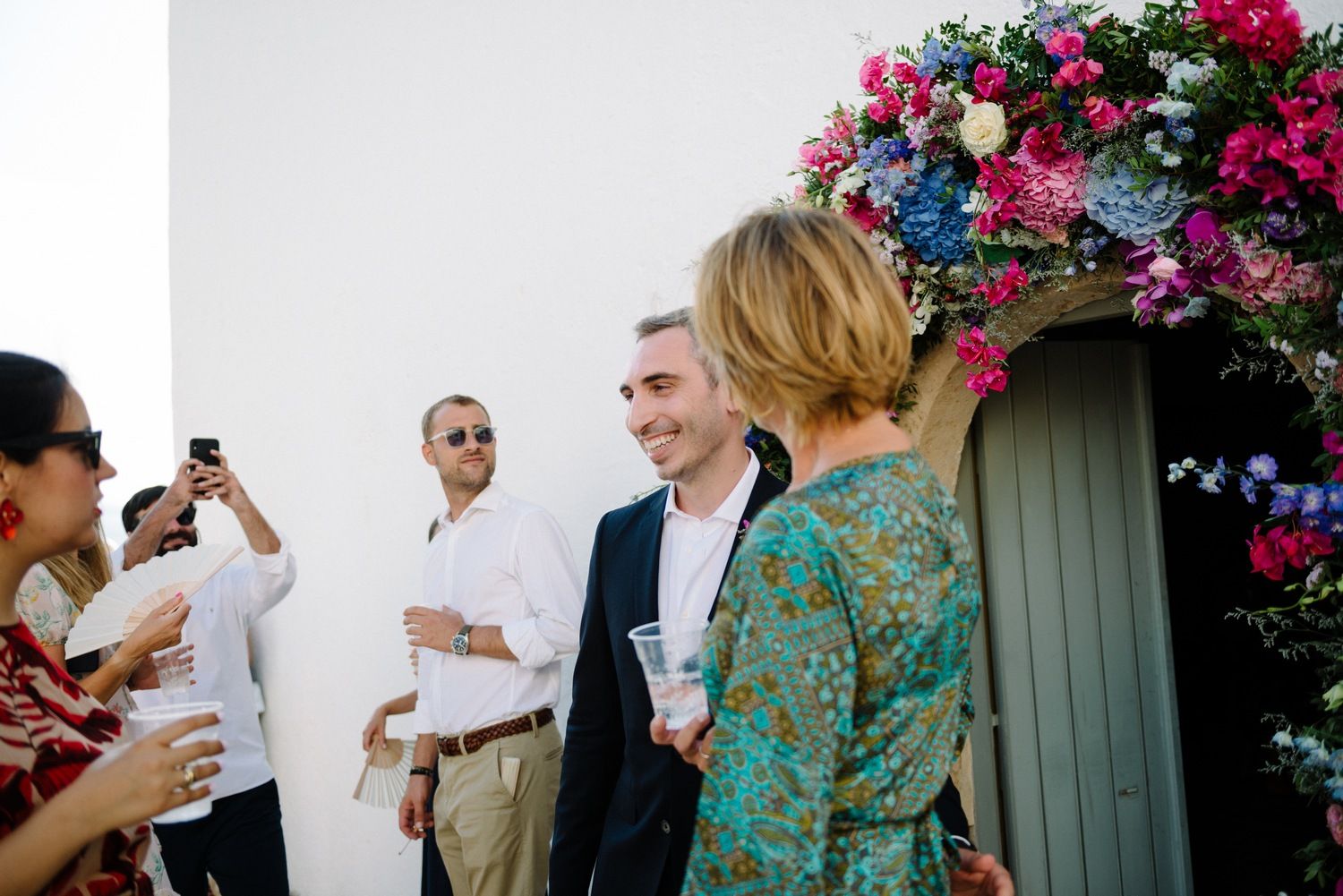 Wedding guests gathered around vibrant floral archway during outdoor celebration.