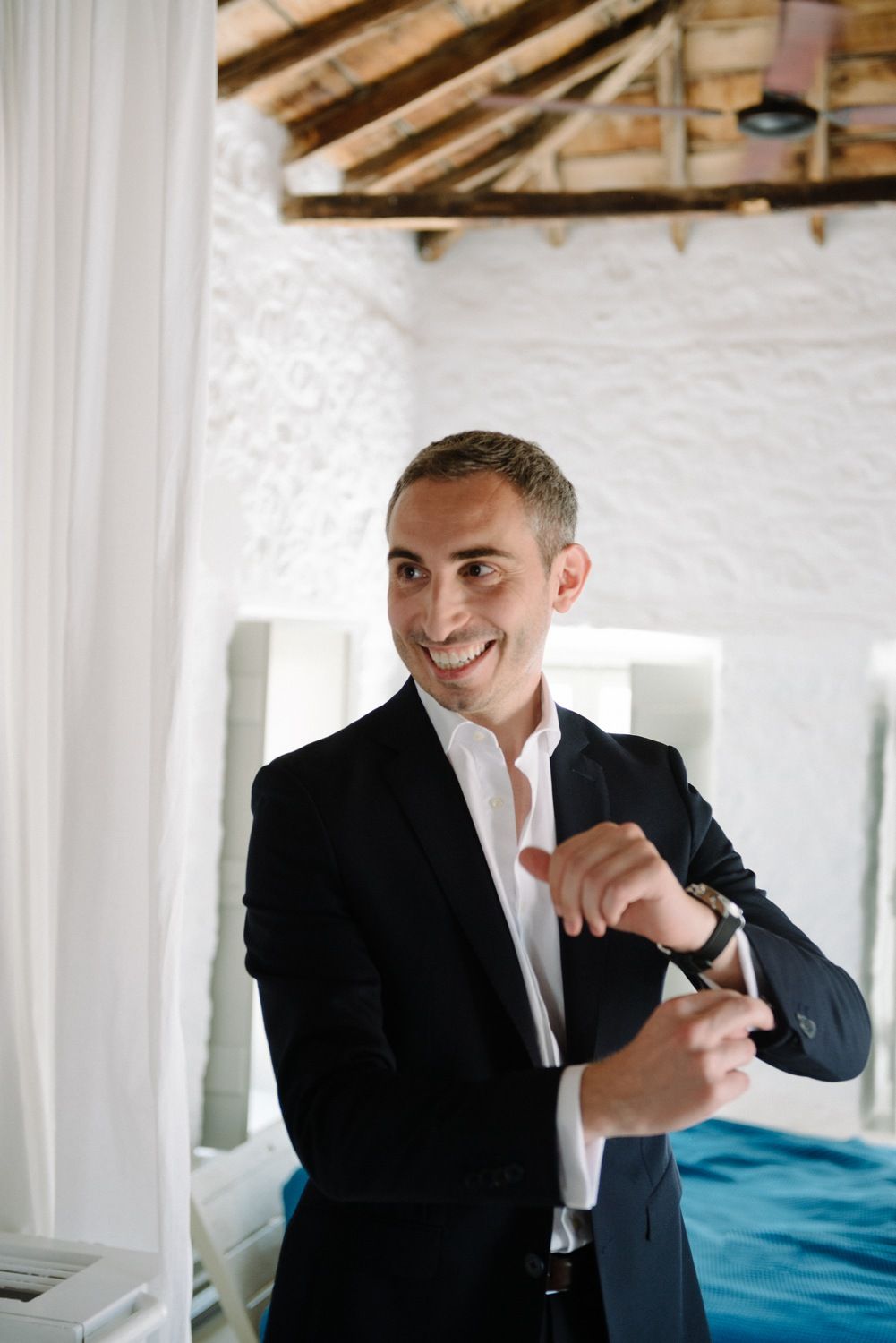 A man wearing a navy suit adjusts his cufflinks while smiling in a white room.