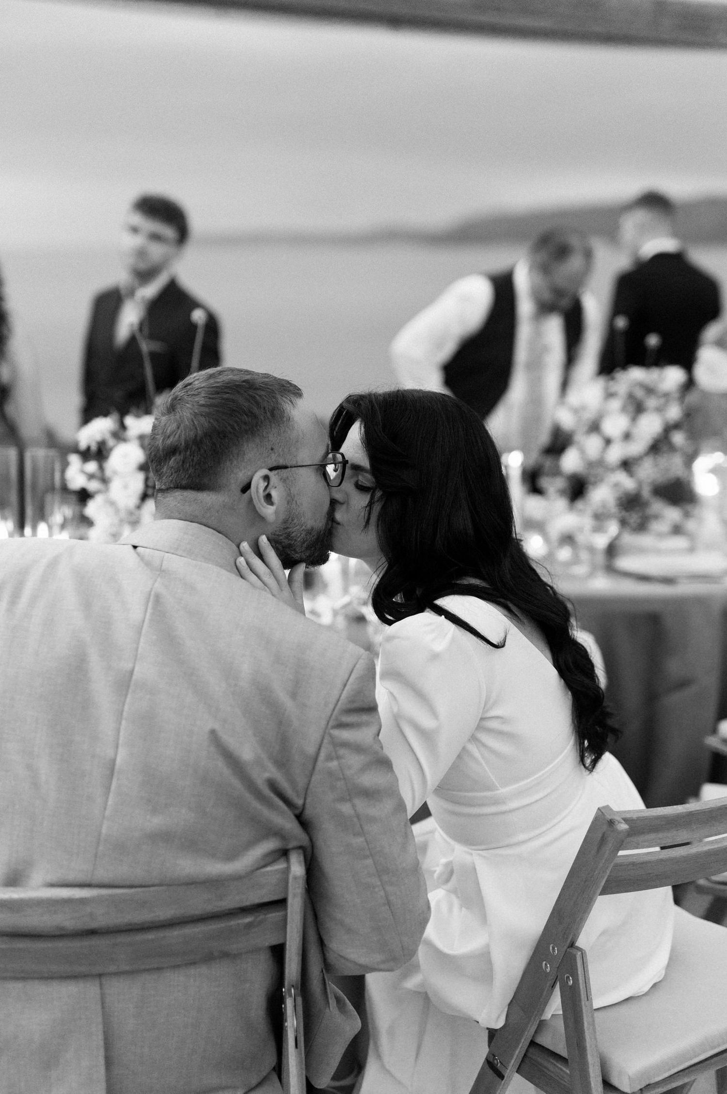 A couple shares a romantic kiss while sitting at an elegant wedding reception table with white floral decorations.