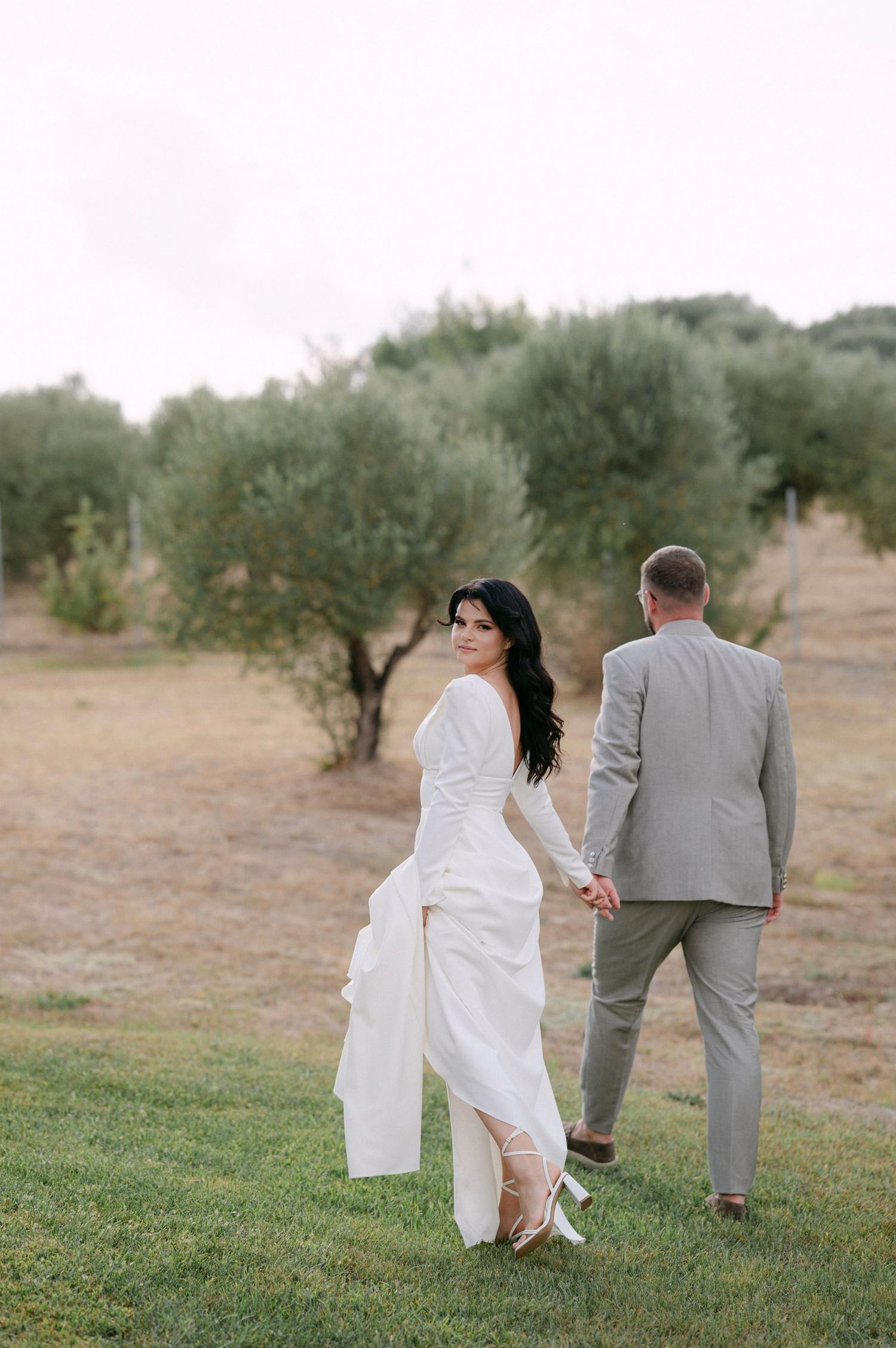 A couple walks together through an olive grove on their wedding day, with the bride in white and groom in gray.