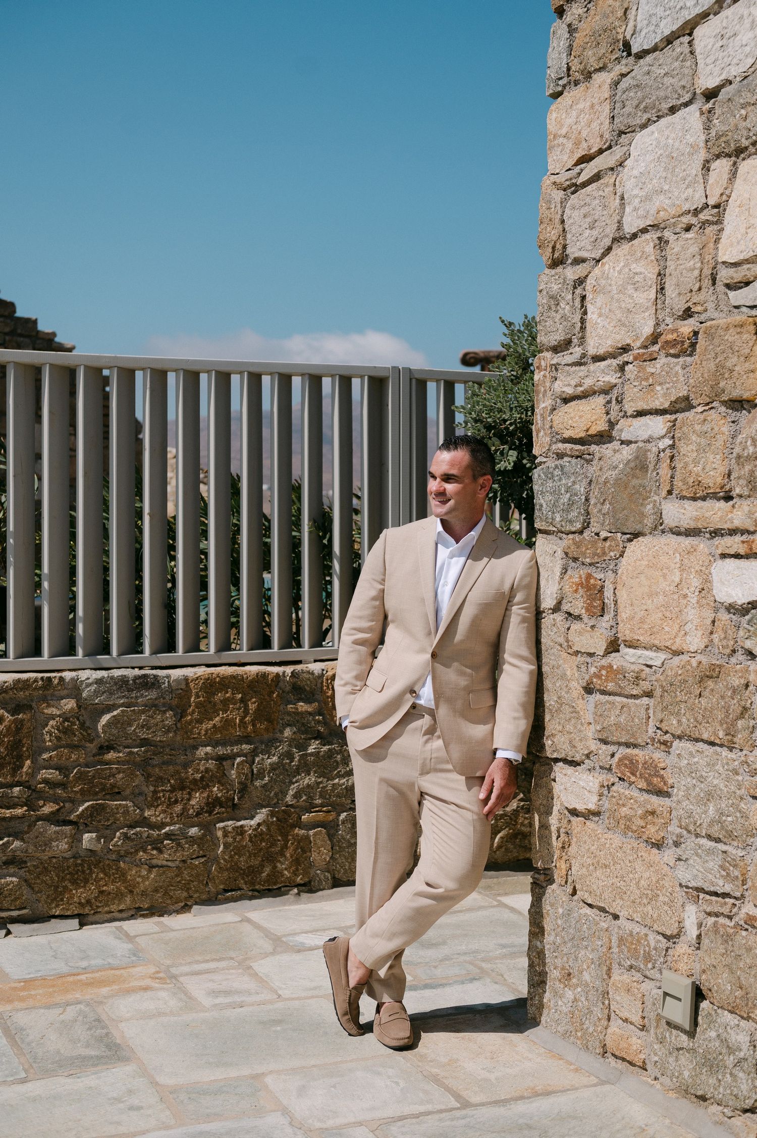Someone in a beige suit sits on stone steps against a rustic wall with vertical metal railings in the background.