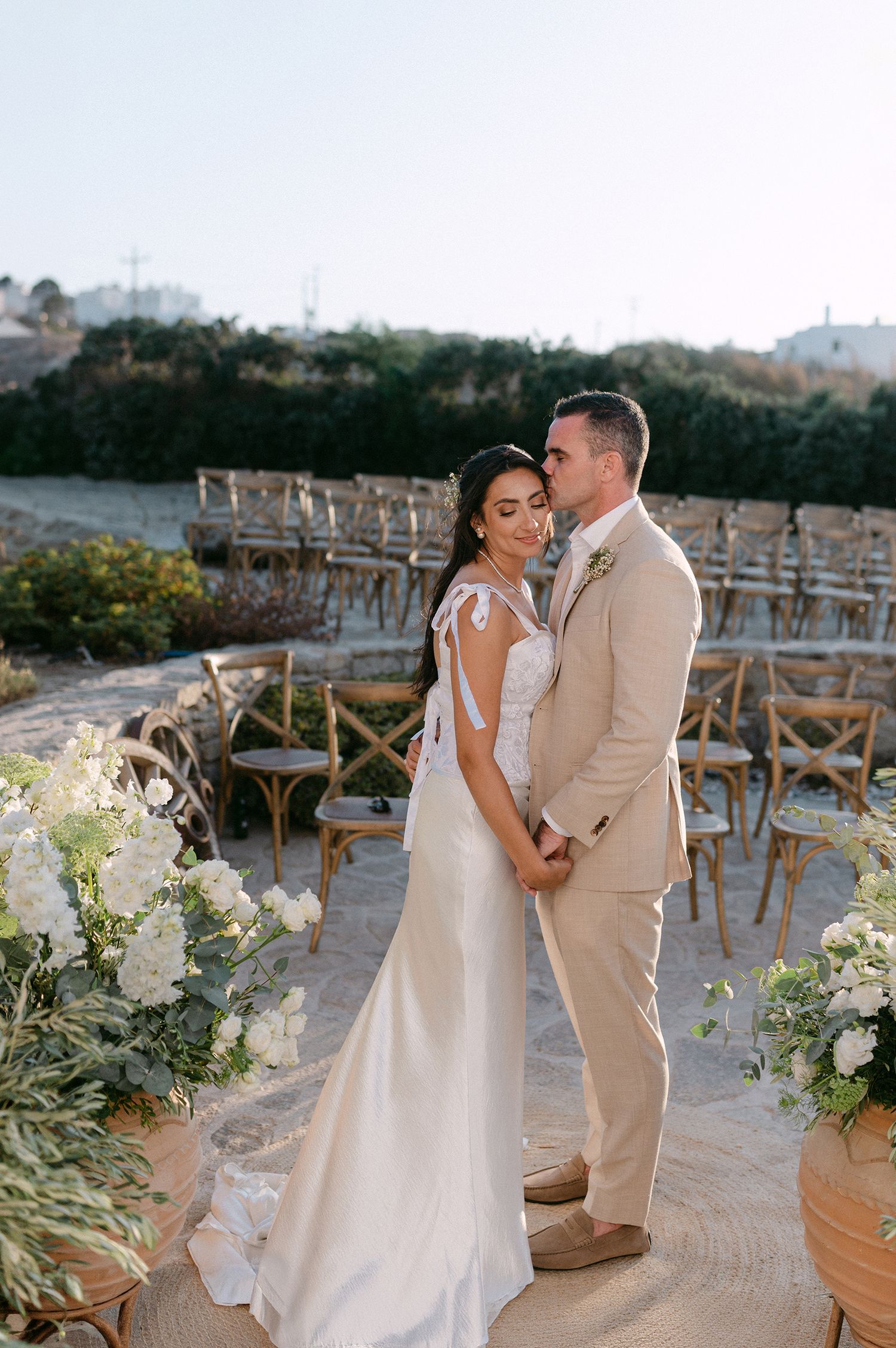 A couple shares an intimate moment at their outdoor beach wedding ceremony with rustic wooden chairs and white floral decor.