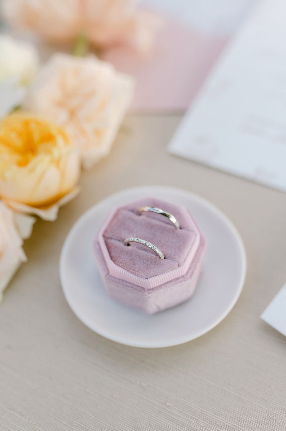 Pink hexagonal ring box with delicate wedding band displayed on white plate with peach roses in background.