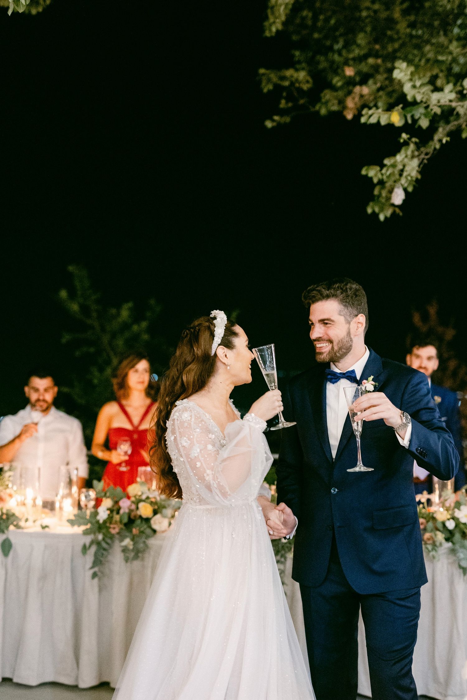 A wedding couple share a celebratory toast at their outdoor reception with guests and floral decor in the background.