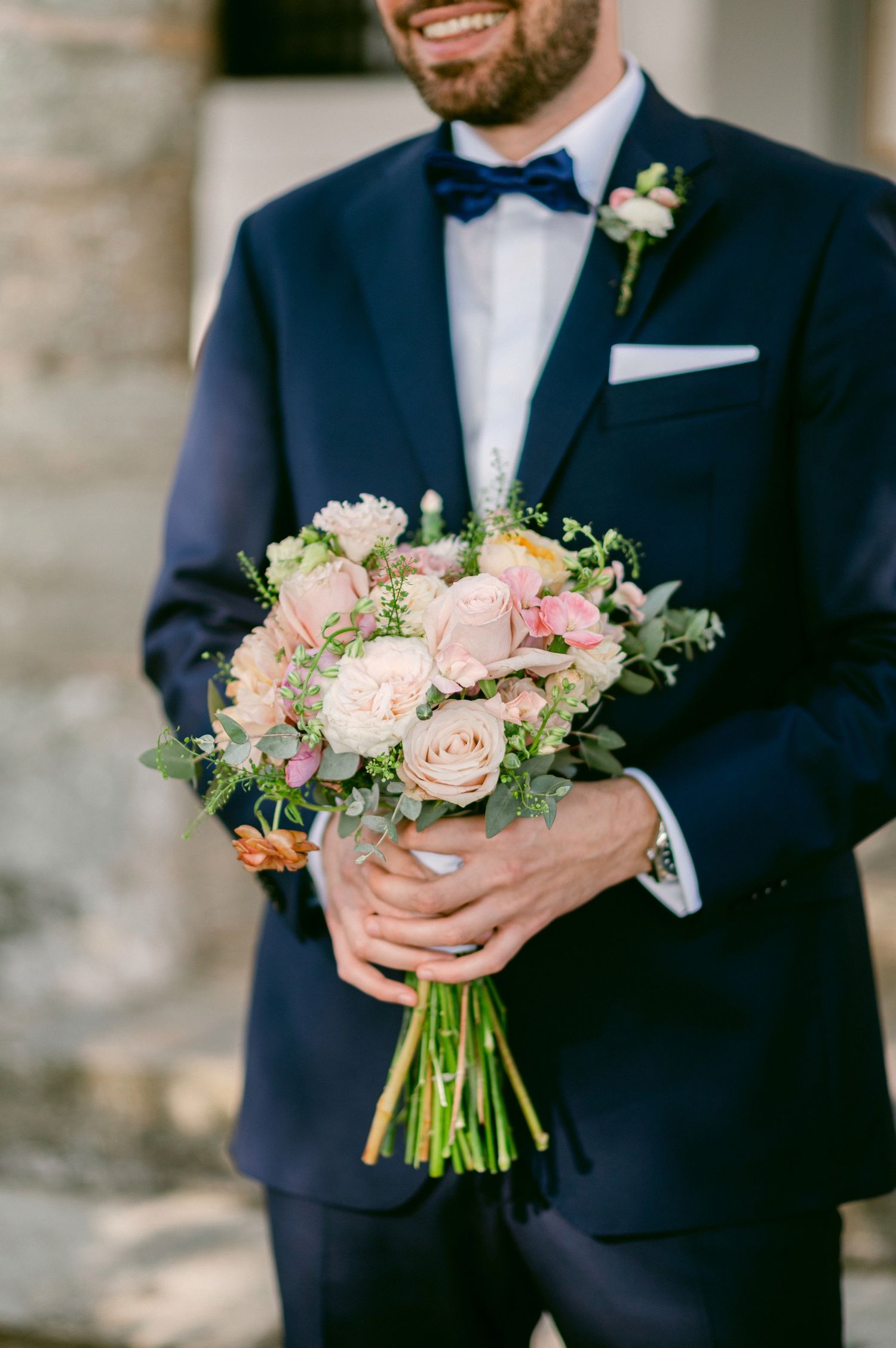 Wedding guest in navy suit and bow tie holds elegant bouquet of blush pink roses and greenery against stone wall.