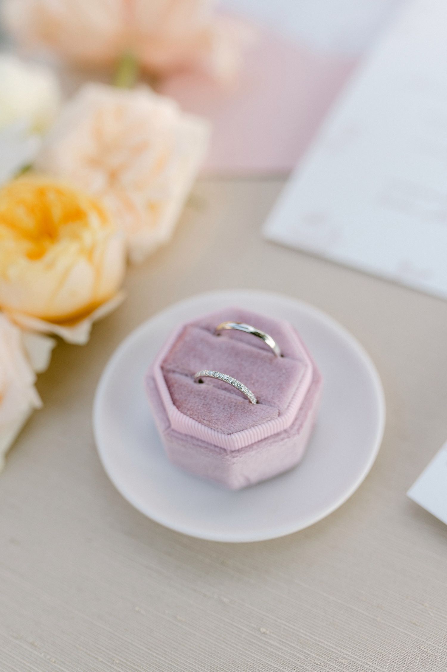 Silver wedding rings resting in a pink velvet octagonal ring box with peach roses in background.