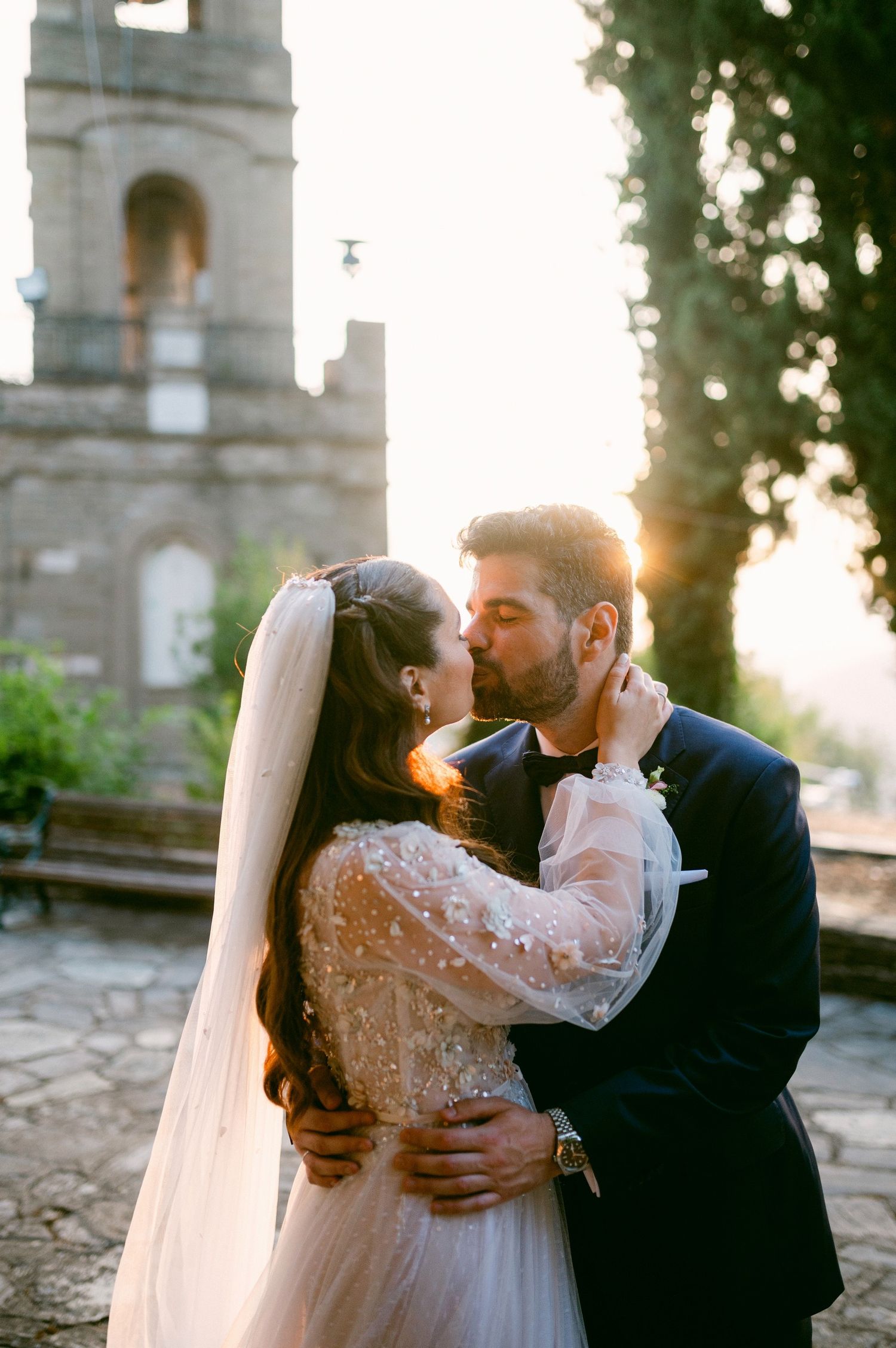 Bride and groom share romantic kiss at sunset in front of historic church tower during golden hour.