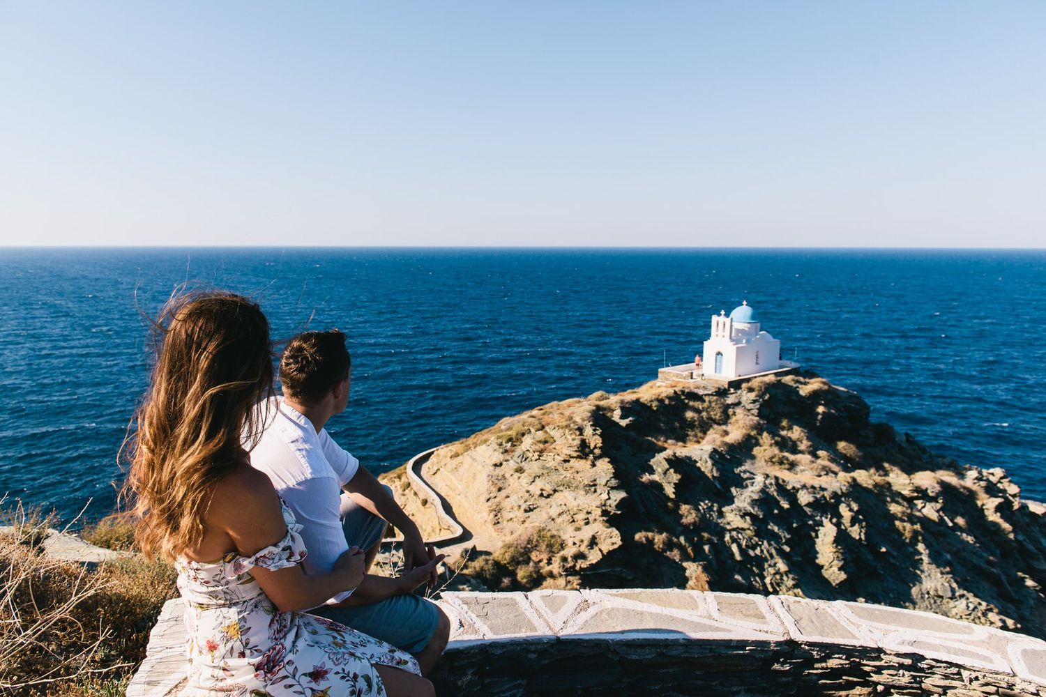 Two visitors admiring scenic white church on cliff overlooking vast blue ocean waters.