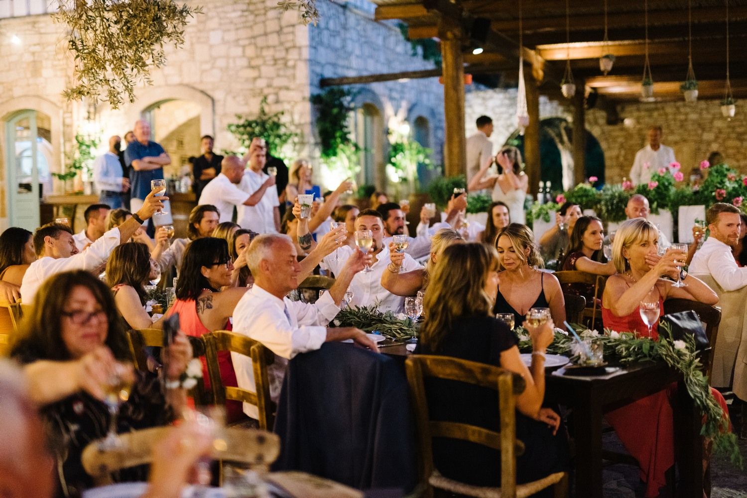 Lively outdoor wedding reception with guests dining under stone arches and ambient lighting.